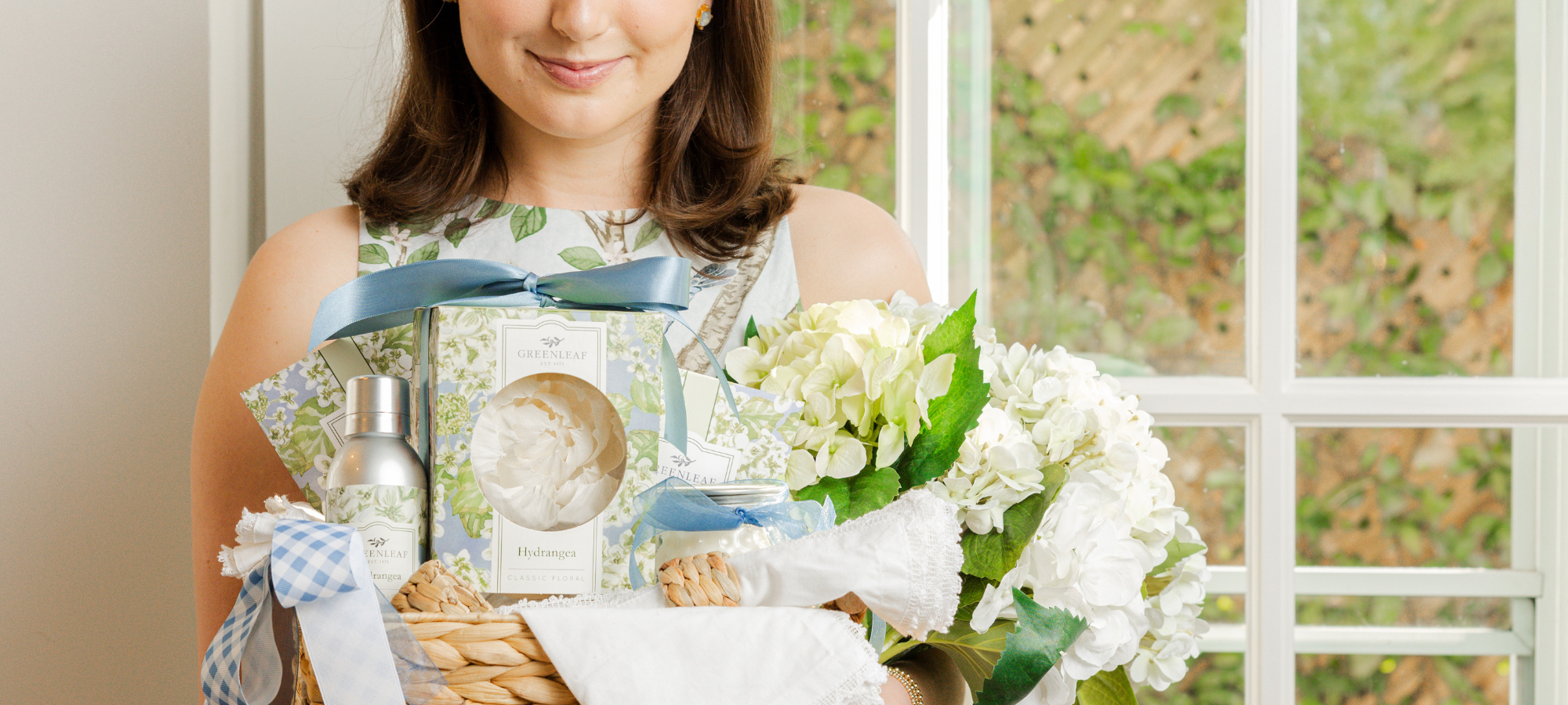 A brunette woman in a dress holding a gift basket of Greenleaf Hydrangea products and hydrangea flowers in front of a bright window