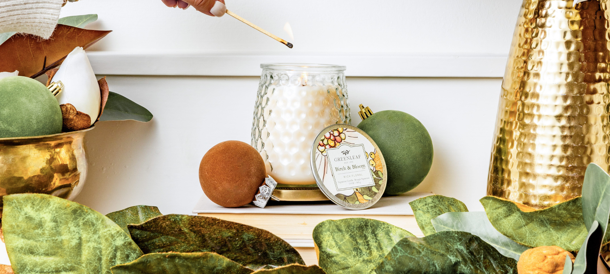 A hand with a match lighting a Greenleaf Birch and Bloom white candle surrounded by magnolia leaves