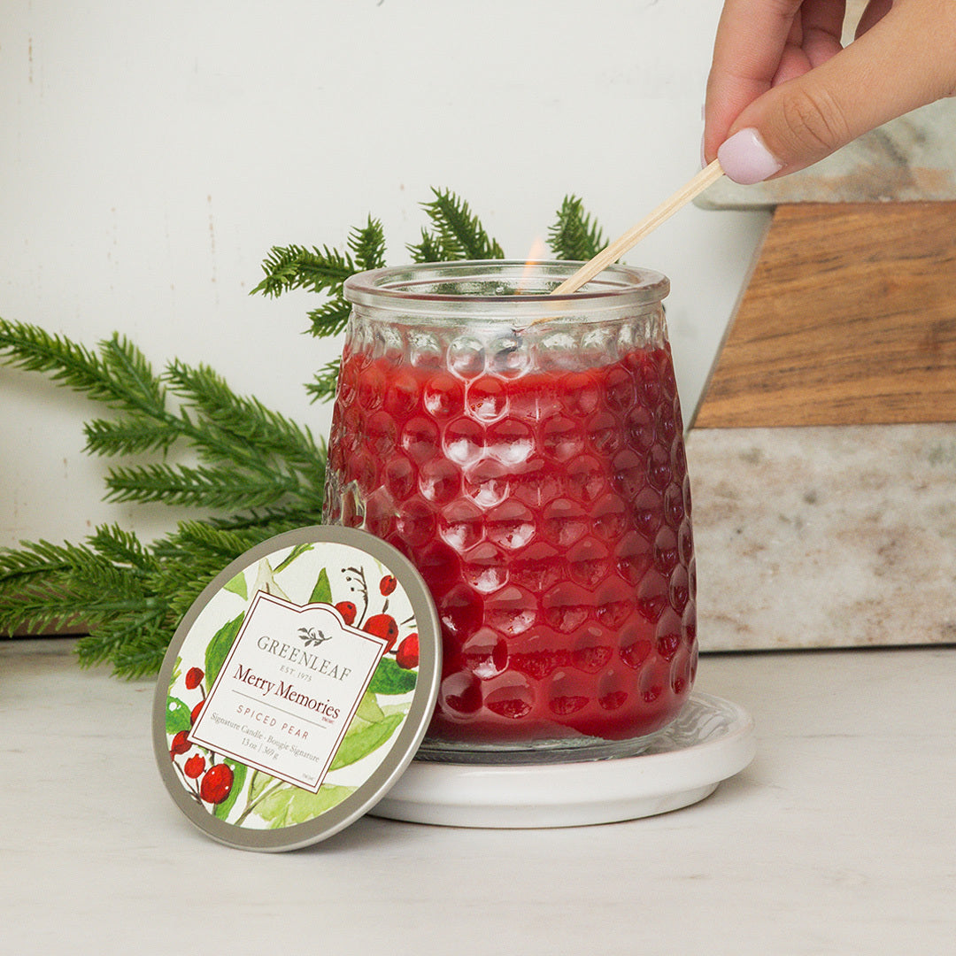 A hand lights a Greenleaf Gifts Merry Memories Signature Candle, its red textured wax on a white coaster with the lid beside it and greenery arranged behind, all set on a white surface.