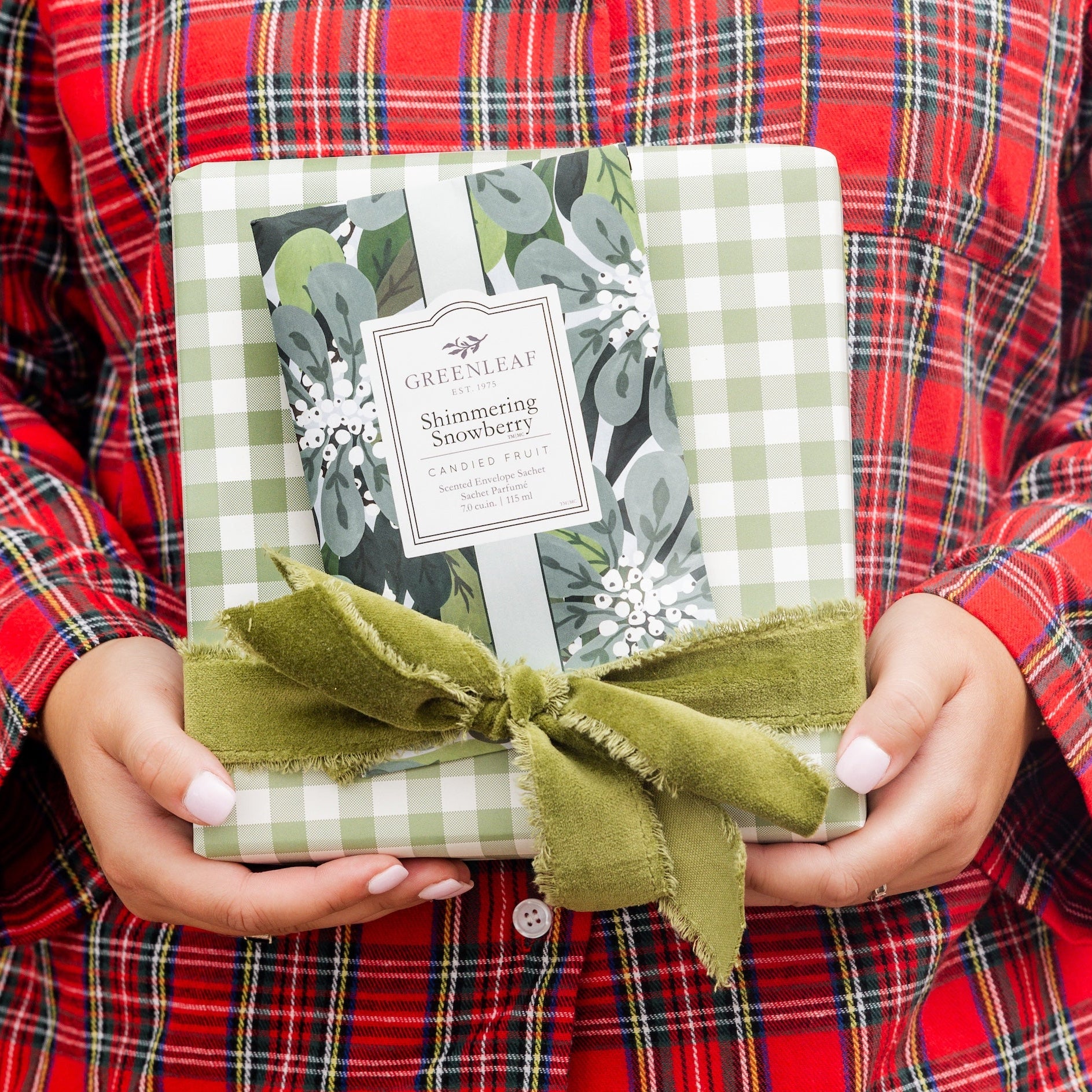 Person wearing a red plaid shirt holding a gift wrapped in green paper with a bow and a Shimmering Snowberry Sachet on top.