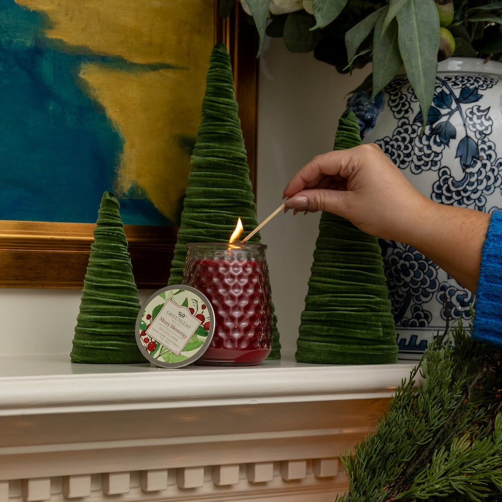 A hand lights a Greenleaf Gifts Merry Memories Signature Candle in a textured red jar on a white mantel, surrounded by green velvet trees and a blue-and-white vase with foliage. The candle’s lid is placed nearby.