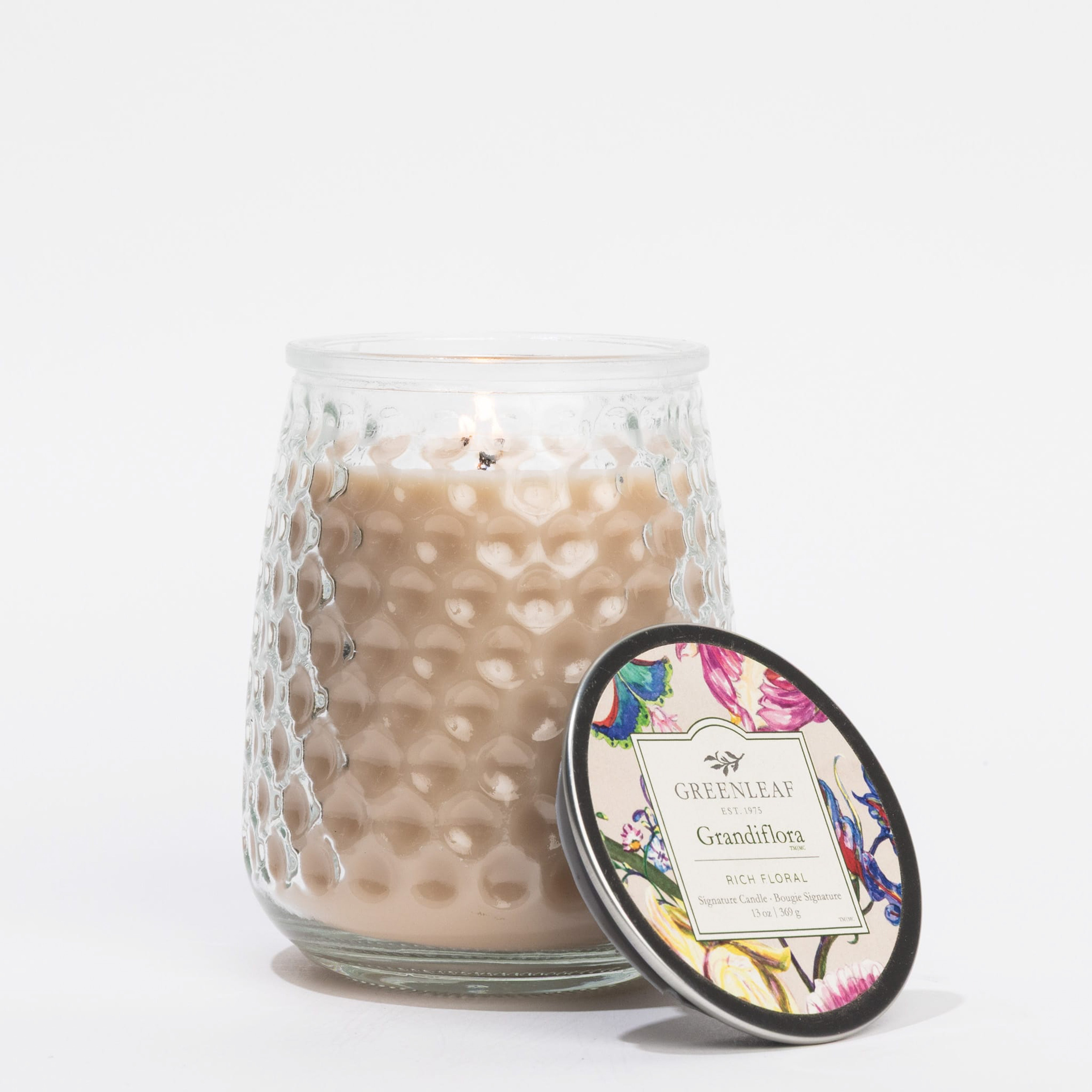 A lit beige Grandiflora Signature Candle by Greenleaf Gifts sits in a textured glass jar with its floral lid resting nearby, the label reading GREENLEAF Grandiflora and showcasing the elegance of Greenleaf fragrances.