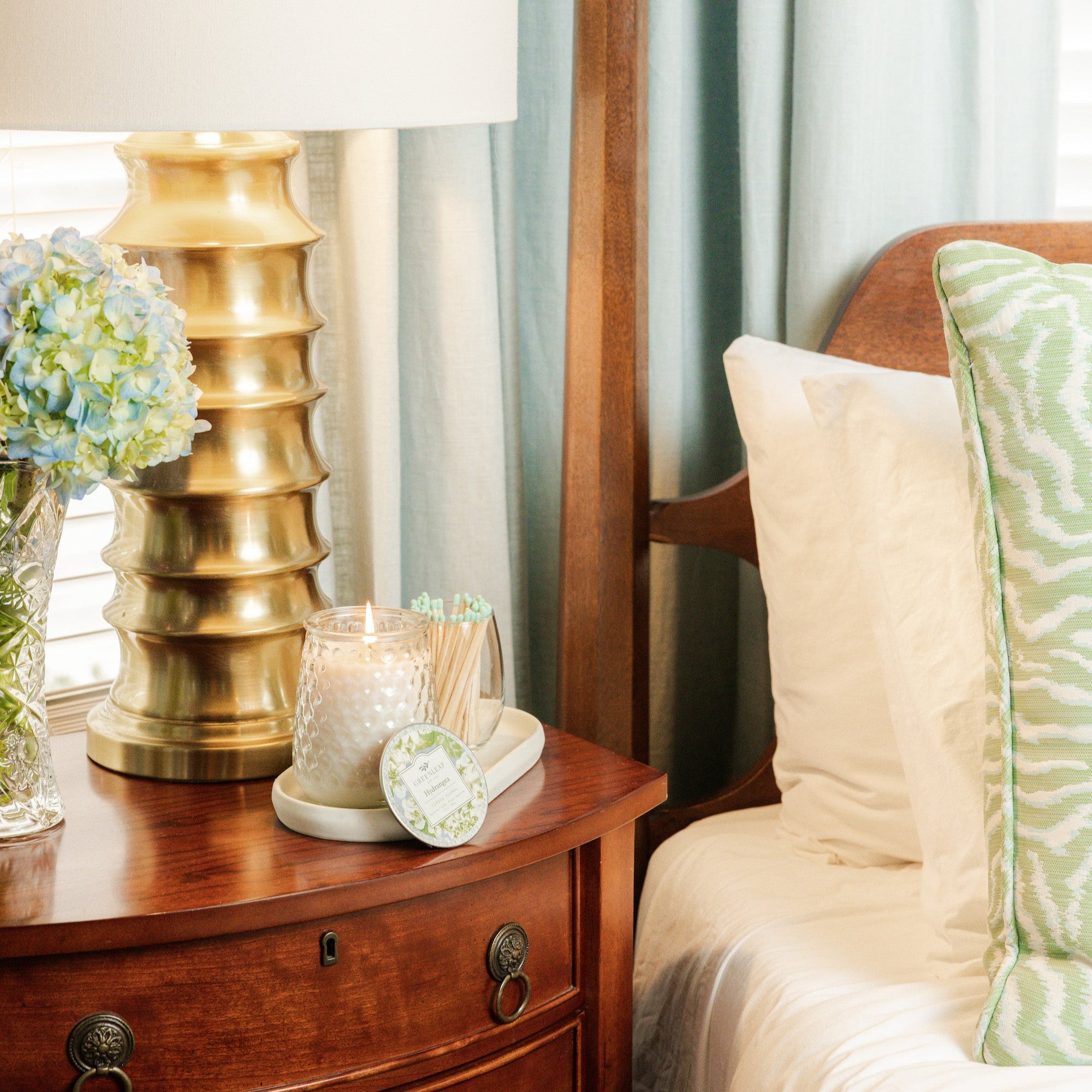 A lit Hydrangea candle on a bedside table with a lamp and flowers