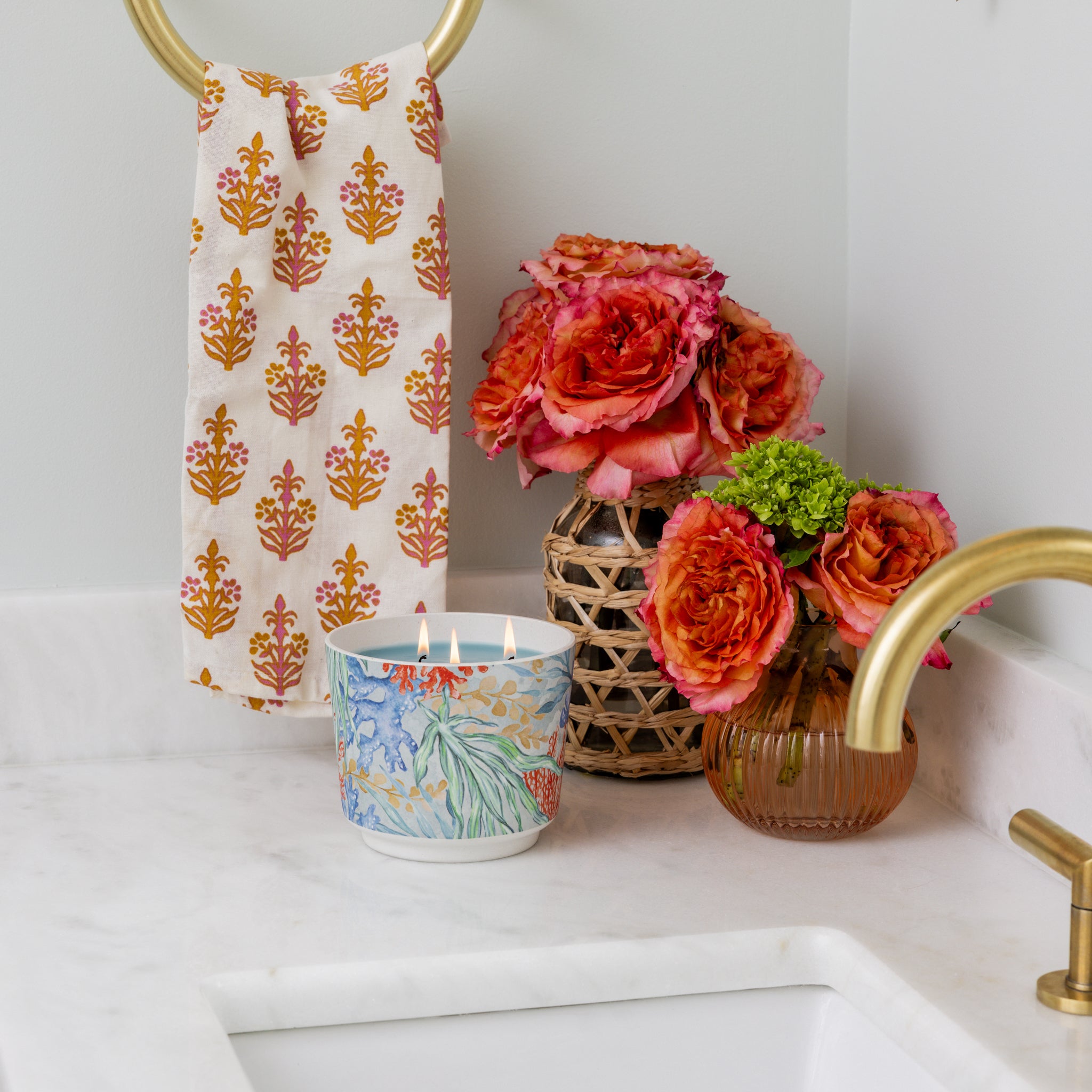 A bathroom countertop features a gold faucet, a white and gold patterned hand towel, the Greenleaf Gifts Seaspray Patterned 3 Wick Candle burning in a decorative holder, and two vases with vibrant orange and pink flowers.