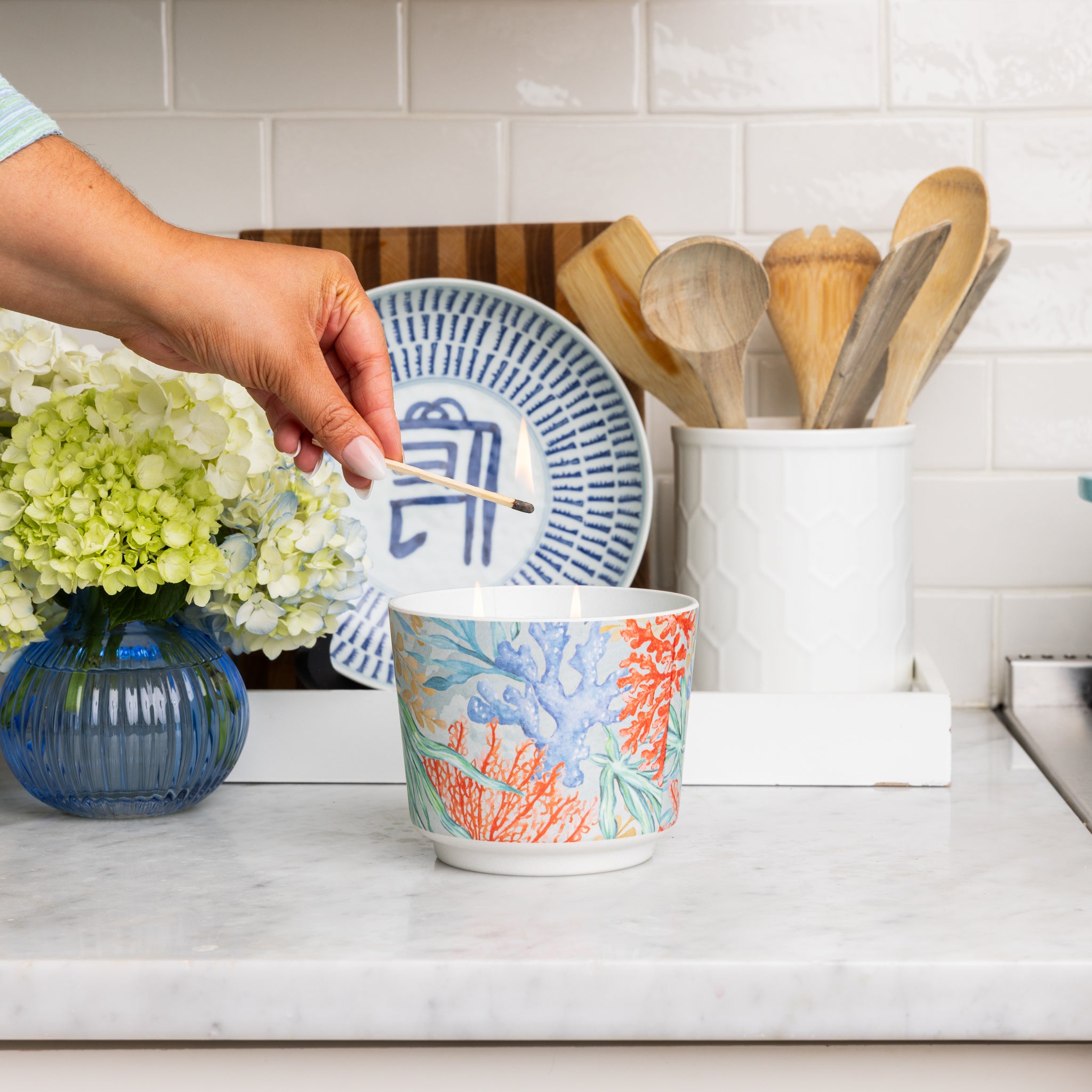 A hand stirs a spoon in a colorful bowl on the kitchen counter, next to Greenleaf Gifts' Seaspray Patterned 3 Wick Candle, a blue vase with flowers, a plate, and bamboo fiber container with wooden utensils against white tile.