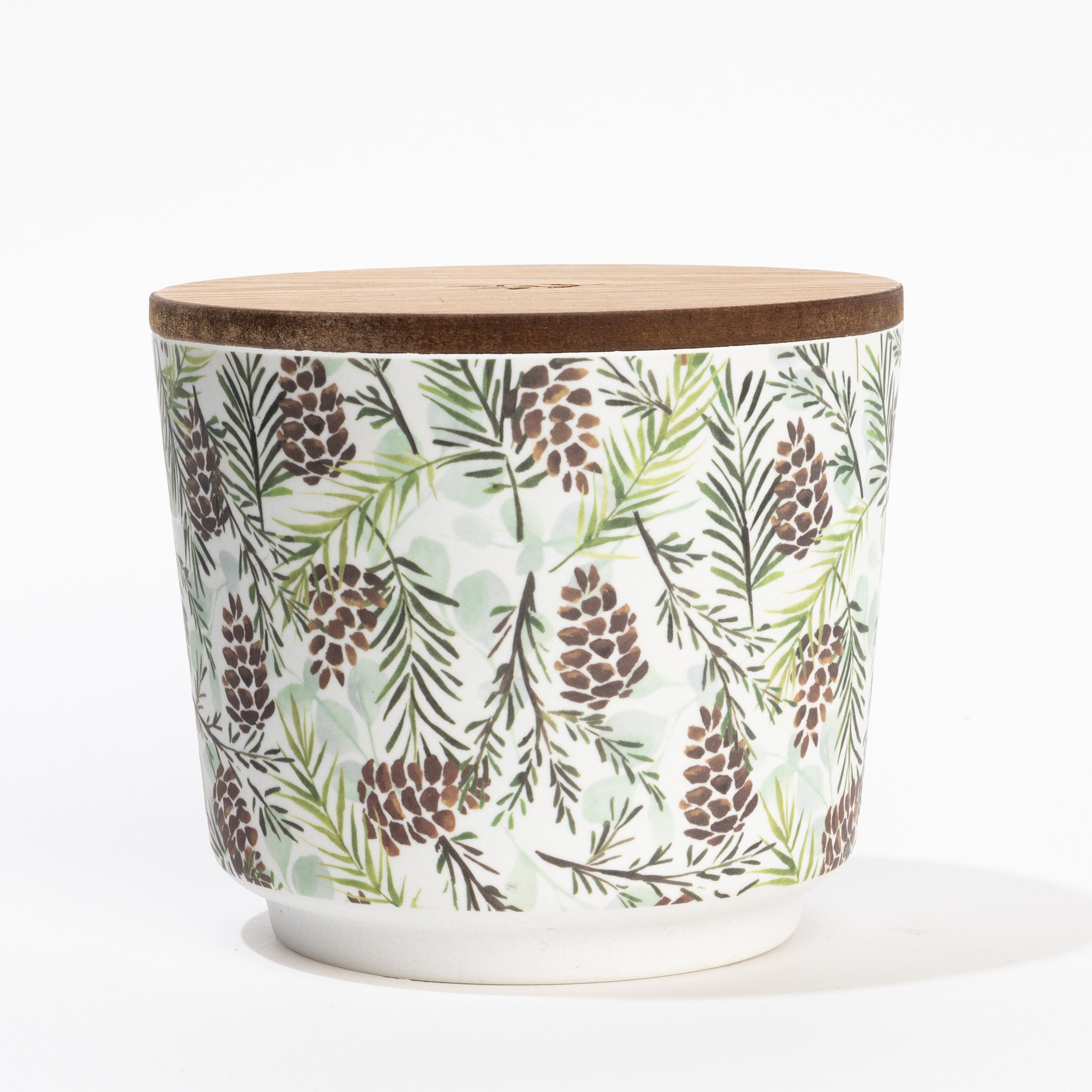 The Greenleaf Gifts Silver Spruce Patterned 3 Wick Candle comes in a white ceramic container with a wooden lid, featuring green pine branches and brown pinecones for a fresh, outdoors-inspired look.