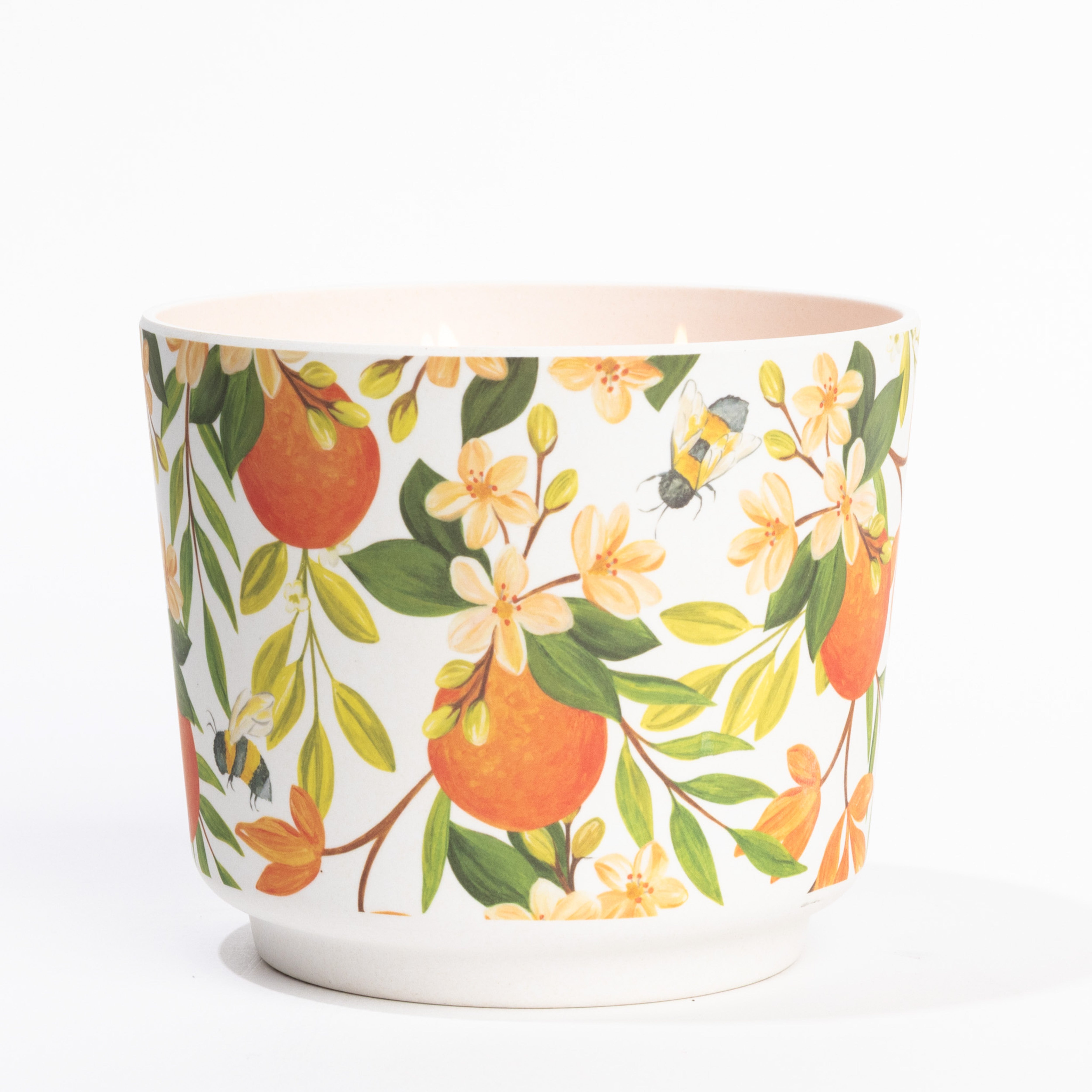 The Patterned 3 Wick Candle - Orange & Honey by Greenleaf Gifts features a vibrant orange, green leaf, blossom, and bee design on white with a light pink interior, reminiscent of its fragrant blend. Shown against a white background.