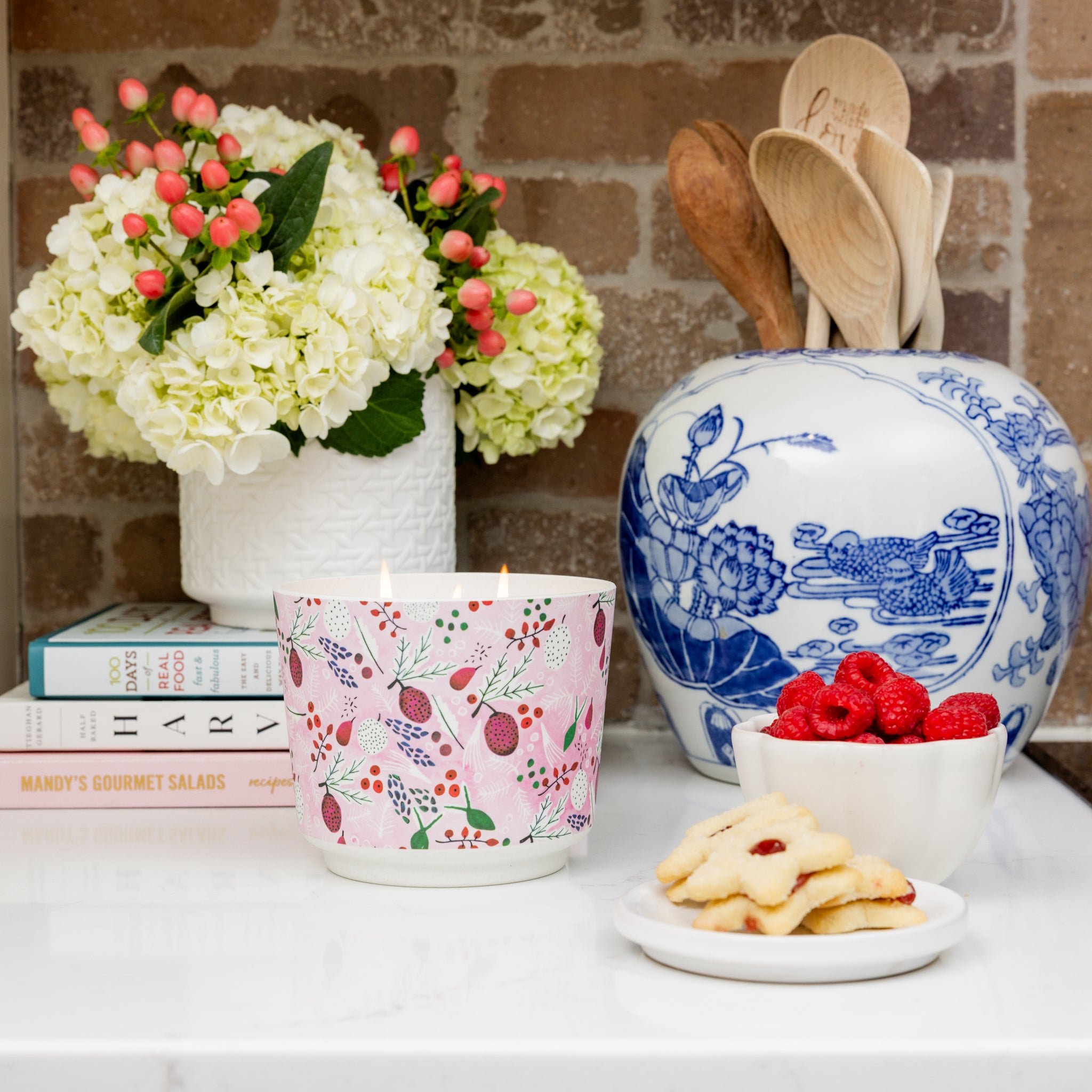 A countertop with a Greenleaf Gifts Brambleberry Patterned 3 Wick Candle, a vase of white flowers, a blue-patterned jar of wooden utensils, stacked cookbooks, berry-topped cookies on a small plate, and a bowl of raspberries.