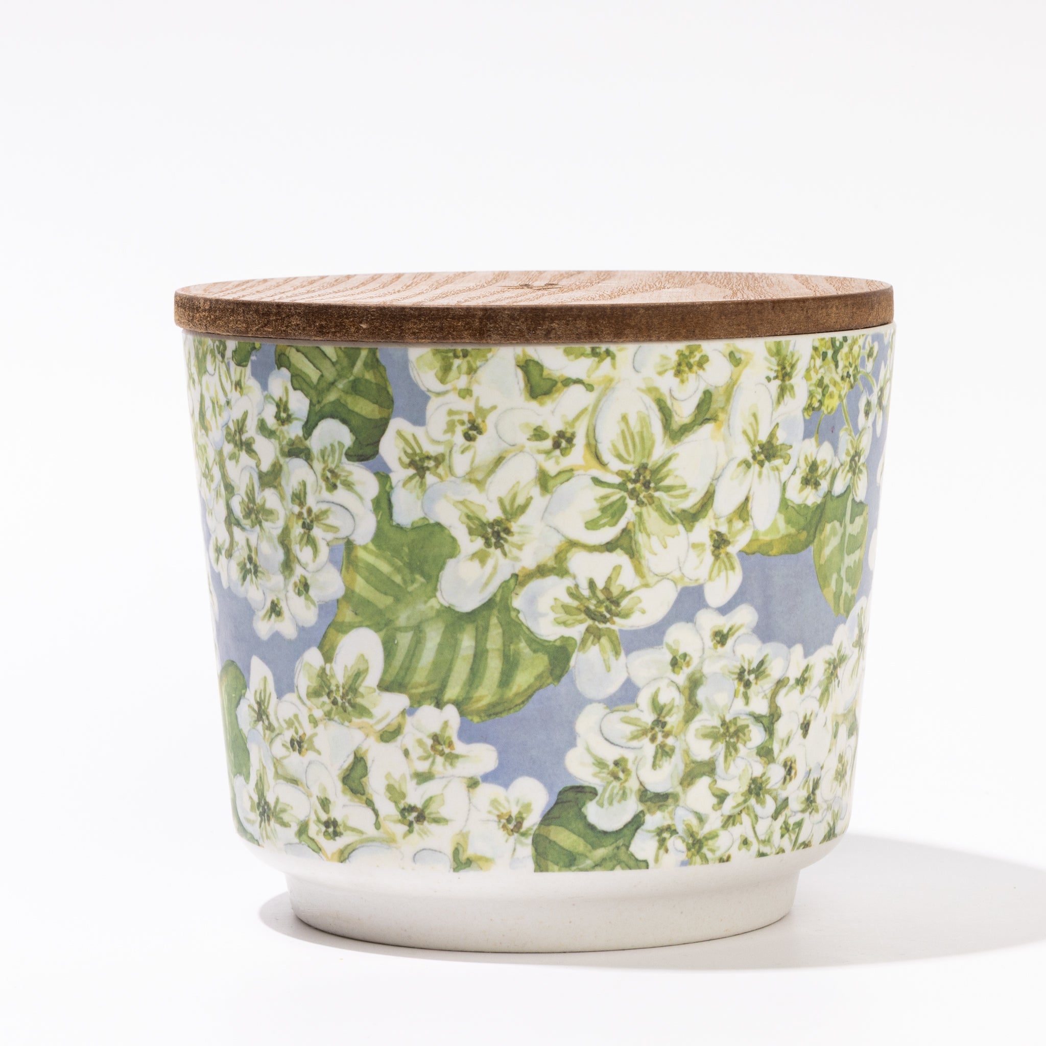 The Hydrangea Patterned 3 Wick Candle by Greenleaf Gifts features a ceramic container with a wooden lid and a white and green floral design on light blue, displayed against a plain white background.