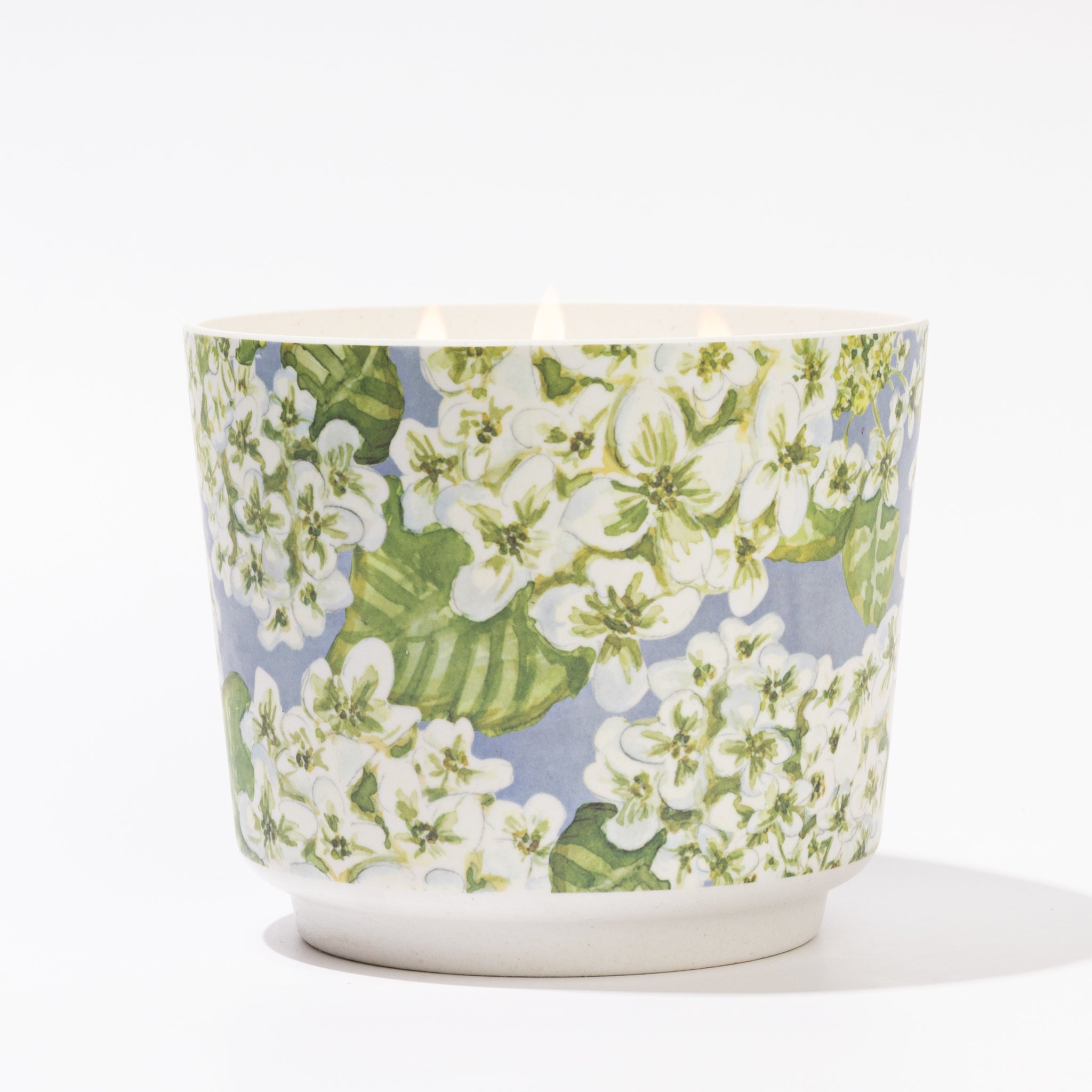 The Hydrangea Patterned 3 Wick Candle by Greenleaf Gifts is a white ceramic holder with a floral hydrangea design of white blossoms and green leaves on light blue, featuring a lit candle with hydrangea fragrance.