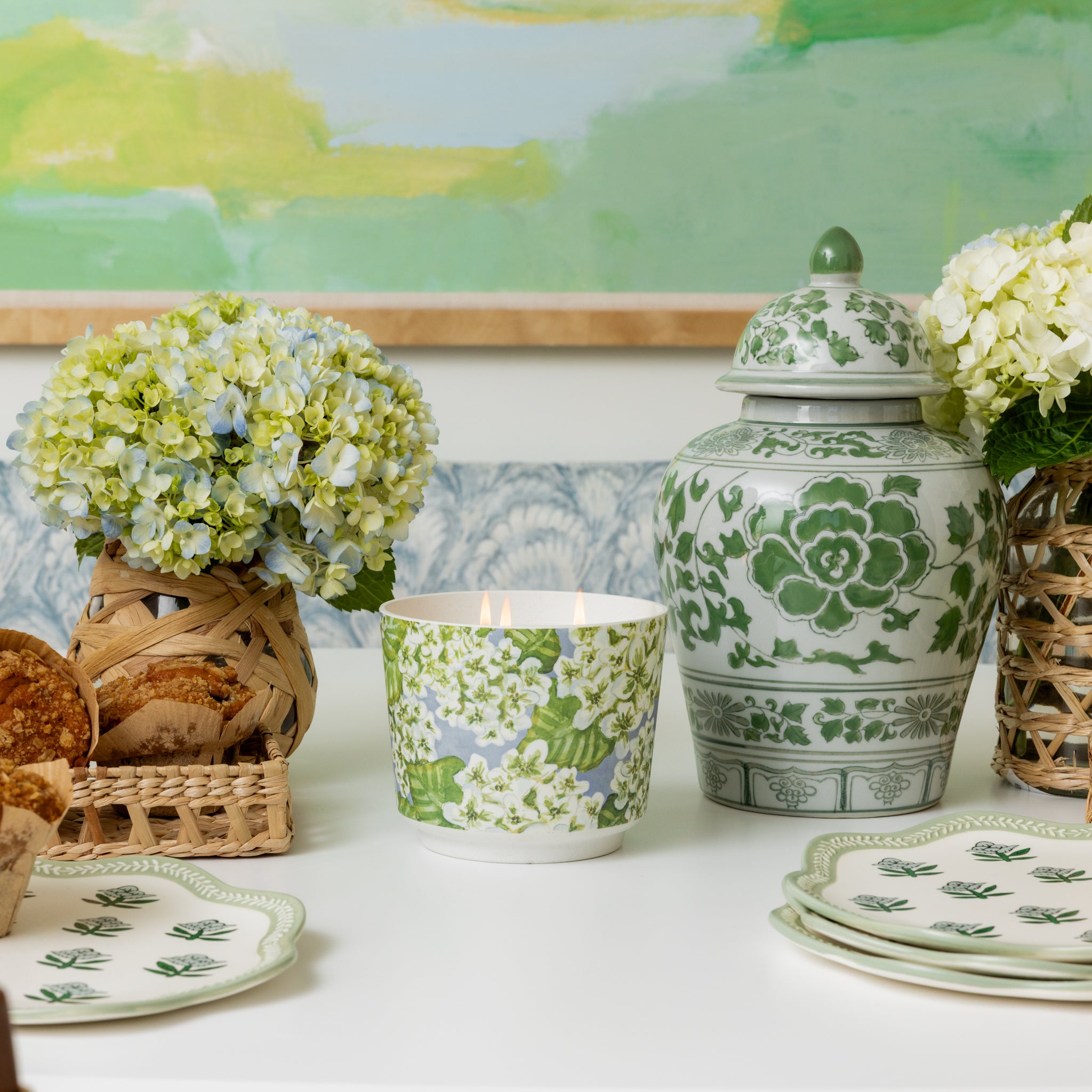 A table features green and white floral china, Greenleaf Gifts’ Hydrangea Patterned 3 Wick Candle, plates, baskets of bread, vases of hydrangeas, and a decorative jar set against a pastel green painting.