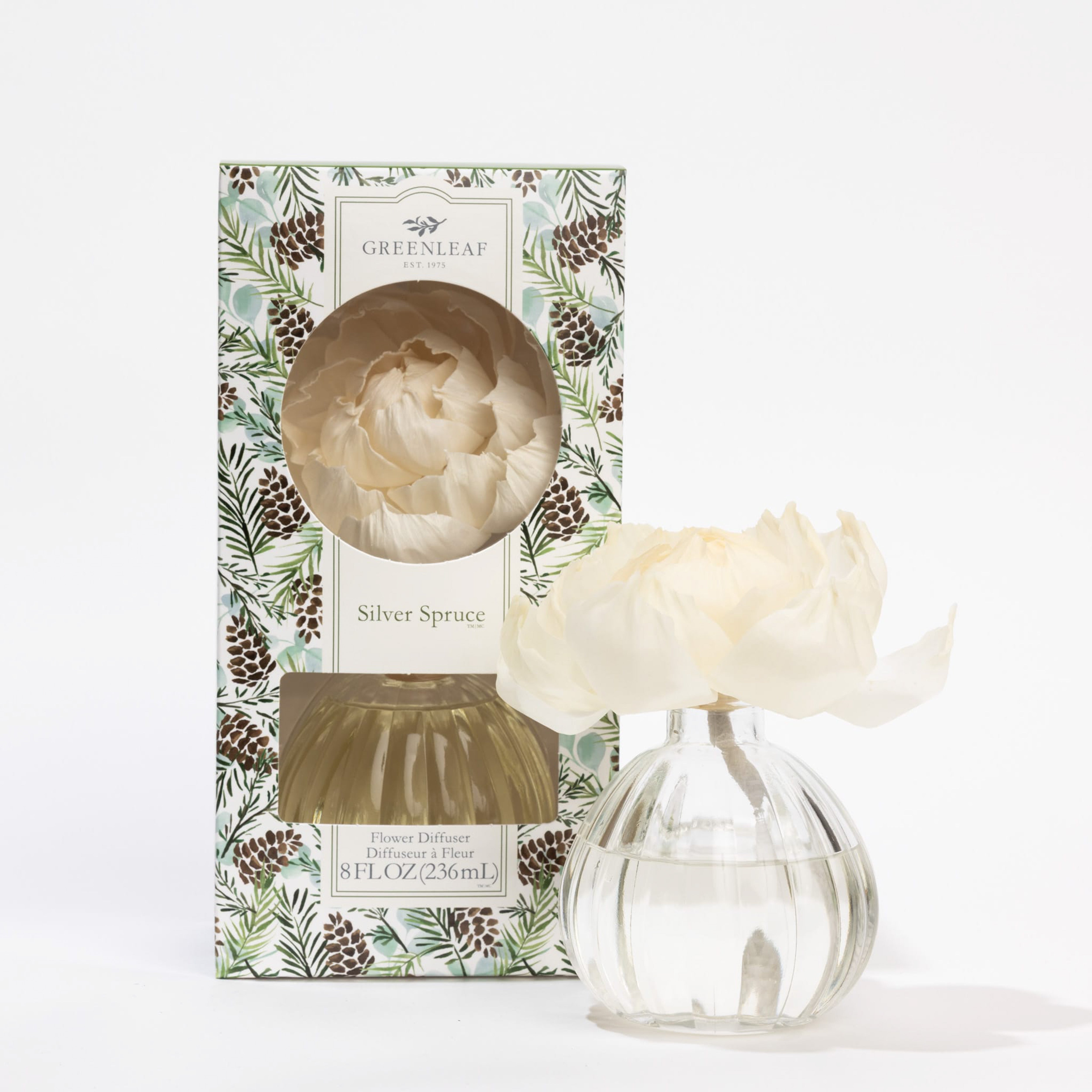 The Greenleaf Gifts Silver Spruce Flower Diffuser features a clear glass bottle with a white artificial flower, filled with scented oil and packaged in a pinecone-and-leaf patterned box.