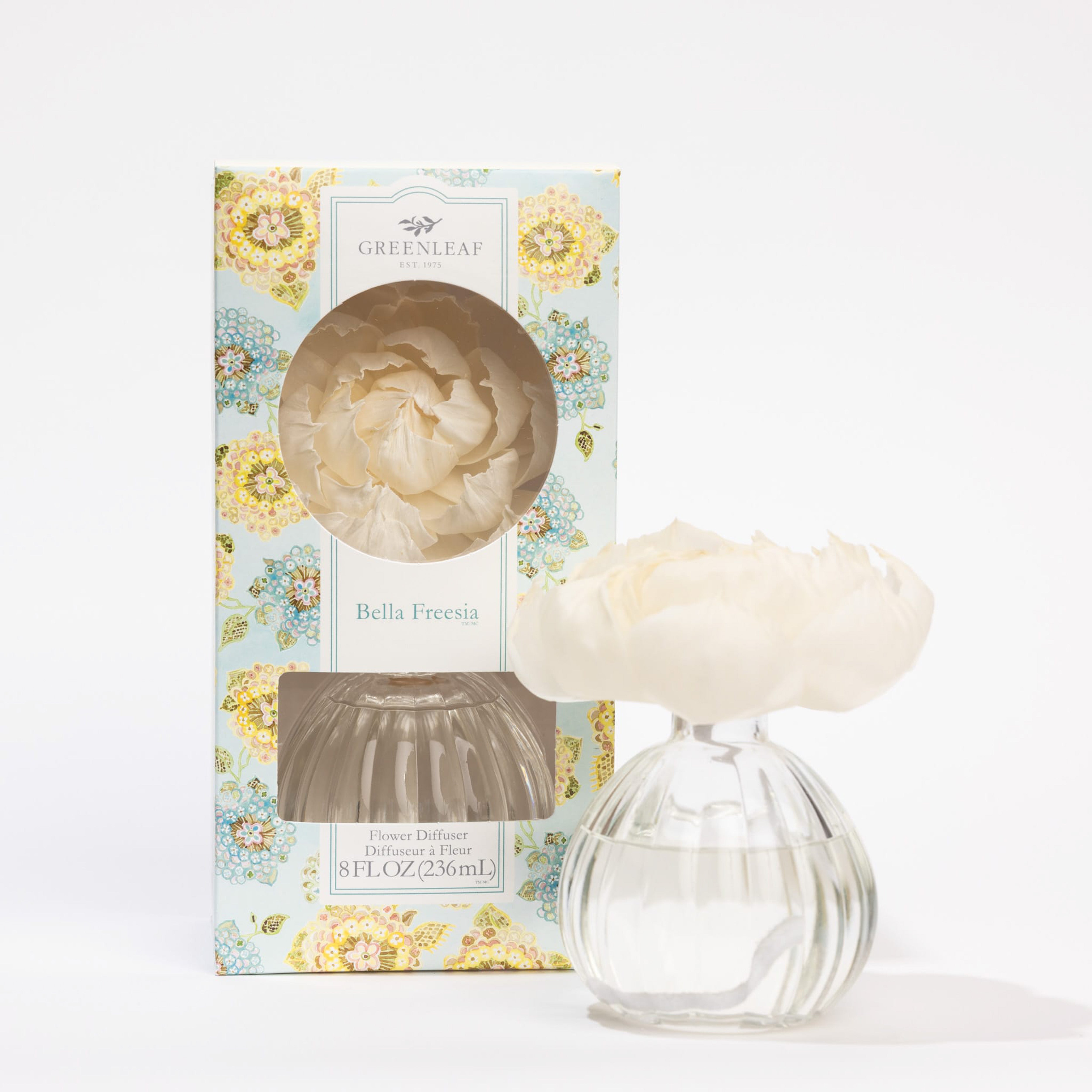 A clear glass vase with a white artificial flower, filled with fragrance oil, stands next to a decorative floral box labeled Greenleaf Gifts Bella Freesia Flower Diffuser with a window showing the matching flower inside.