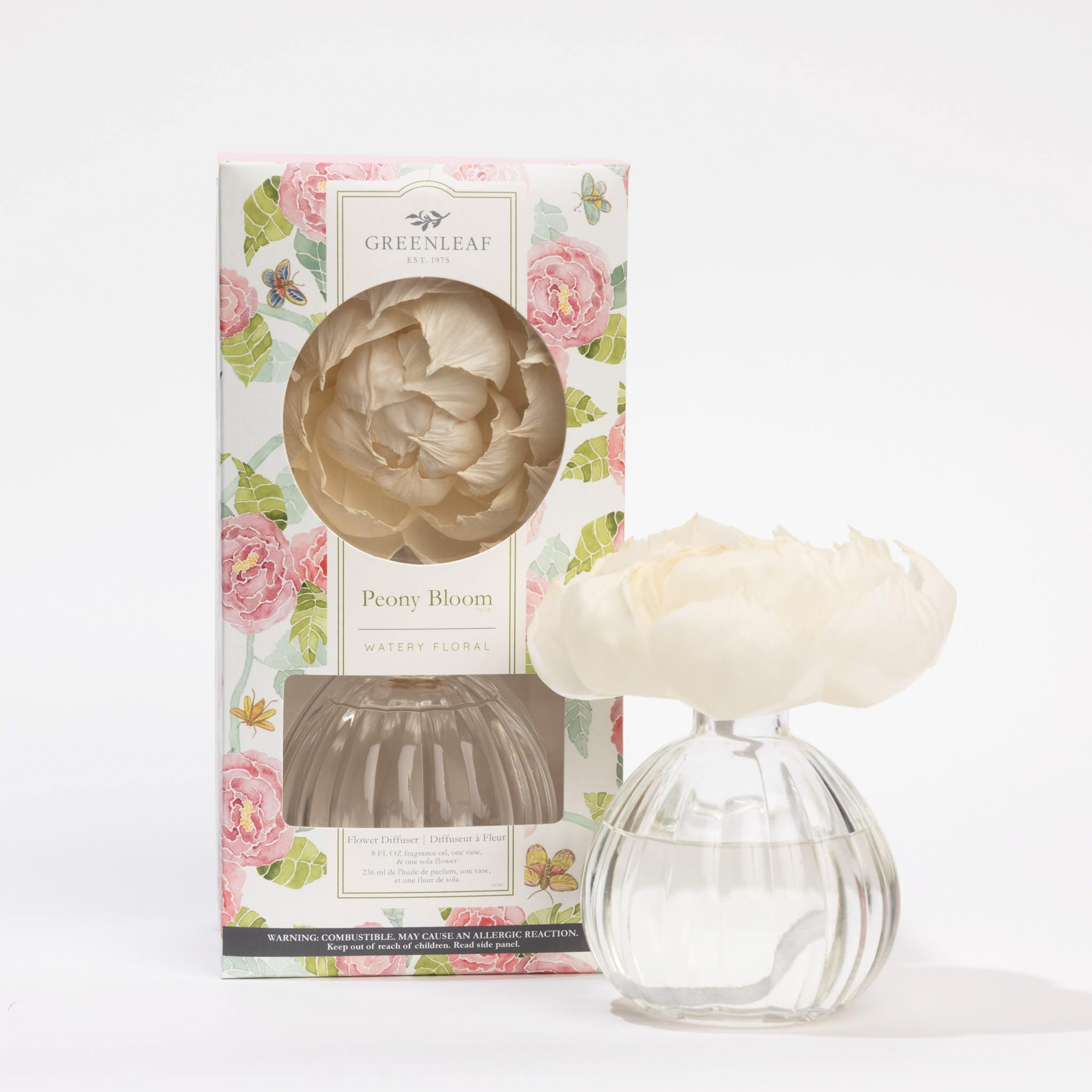 A clear vase filled with fragrance oil holds a white sola paper peony blossom as the Peony Bloom Flower Diffuser by Greenleaf Gifts. It sits beside its decorative floral-patterned box labeled Greenleaf Peony Bloom, Watery Floral.