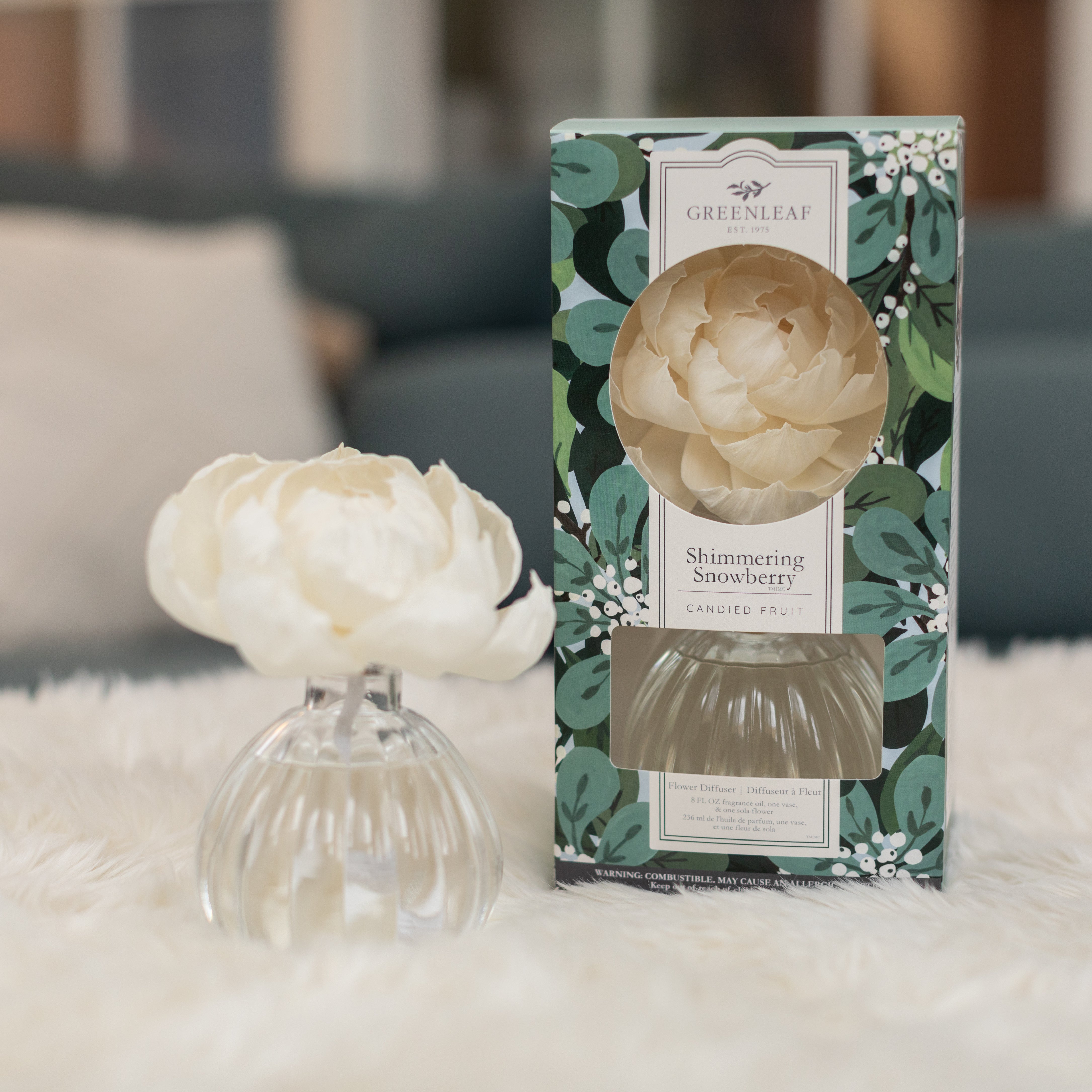 A white artificial flower in a glass vase sits beside a Greenleaf Gifts Shimmering Snowberry Flower Diffuser box with floral and leaf designs, all arranged on a fluffy white surface.