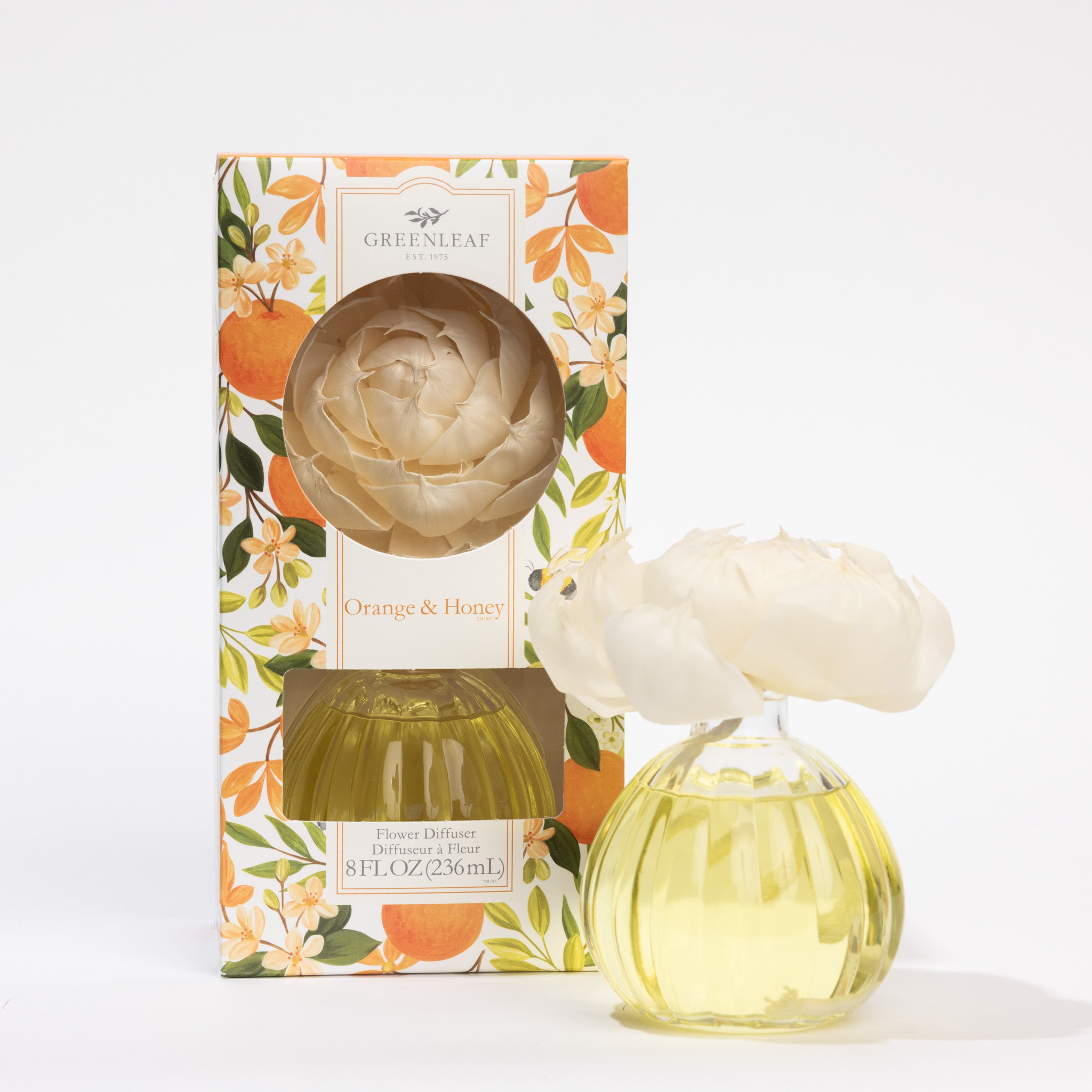 A Greenleaf Gifts Orange & Honey Flower Diffuser with yellow liquid rests next to its floral-themed box, topped with a sola paper flower and adorned in vibrant orange and green accents, all filled with premium fragrance oil.