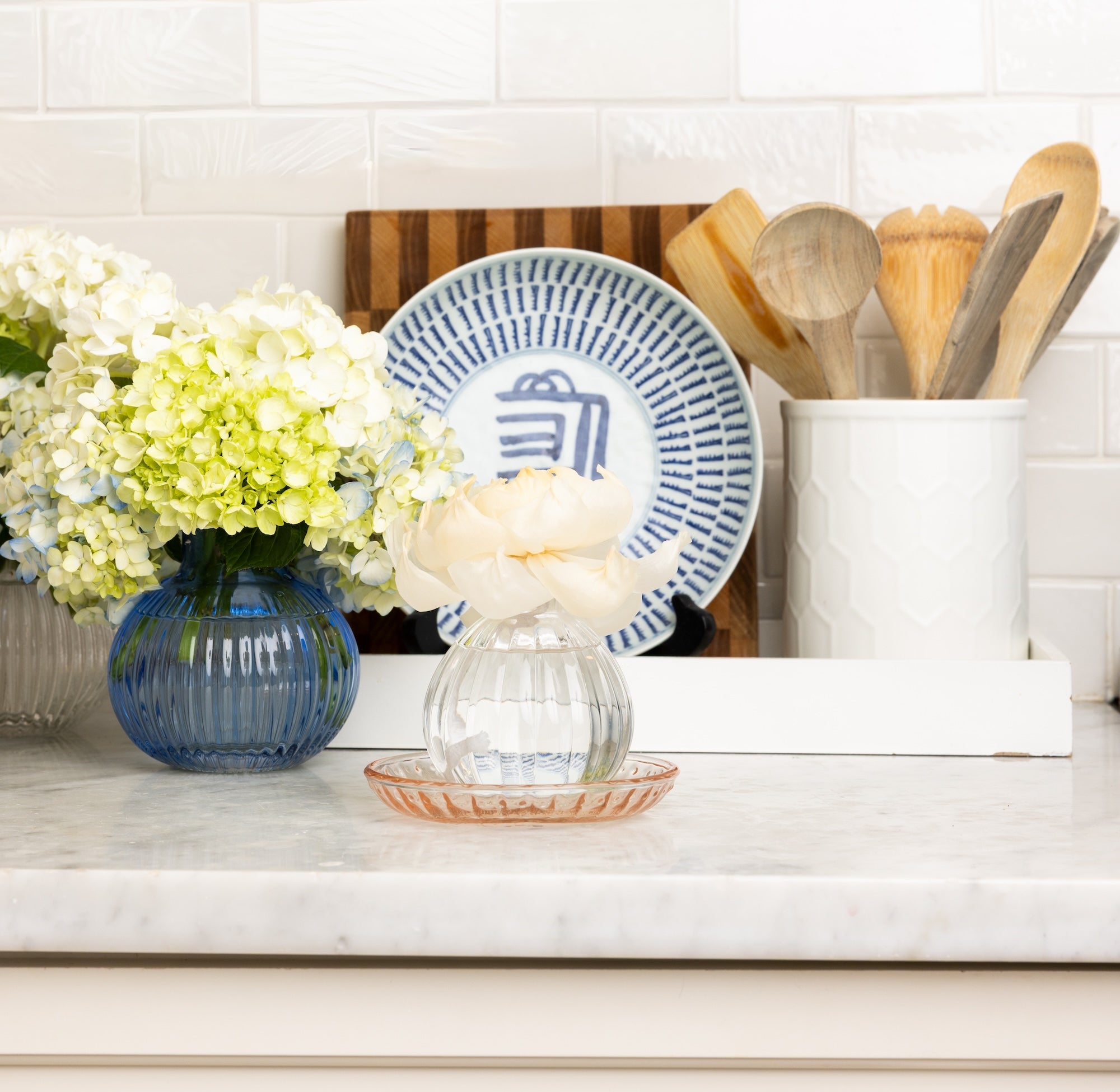A kitchen countertop displays a Greenleaf Gifts Peony Bloom Flower Diffuser, a blue vase of hydrangeas, a decorative blue and white plate, and a white container with wooden utensils.