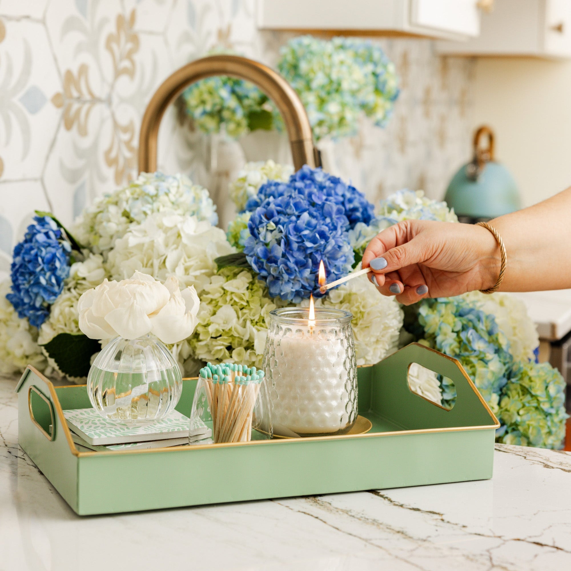 A hand lights a white candle on a green tray beside the Greenleaf Gifts Hydrangea Flower Diffuser and white flowers, with blue and white hydrangeas in the background on a marble countertop.