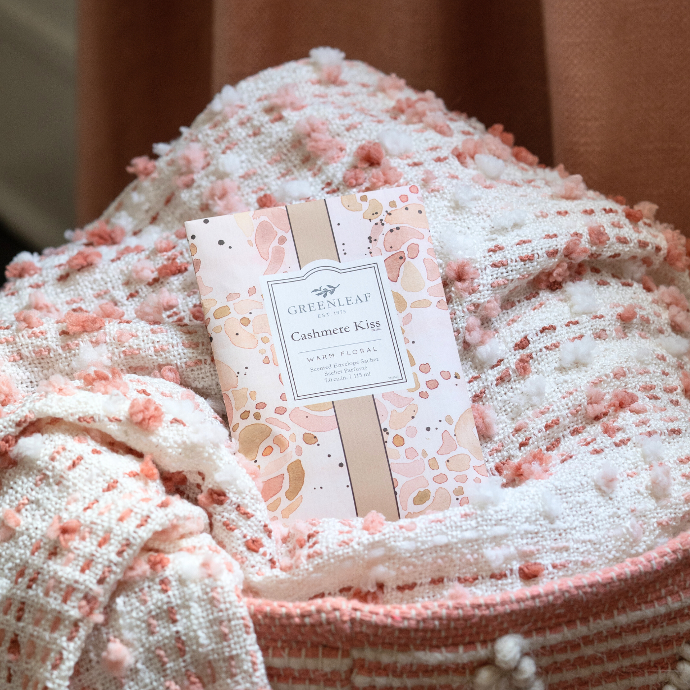 A Greenleaf “Cashmere Kiss” scented sachet rests on a textured white and pink blanket with small pom-poms, next to a peach-colored fabric background.