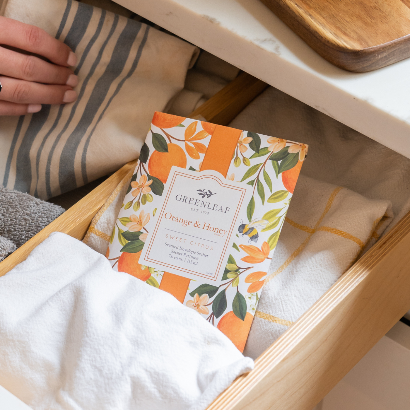 A hand opens a wooden drawer filled with neatly folded towels and a Greenleaf Orange & Honey scented sachet with a floral design.