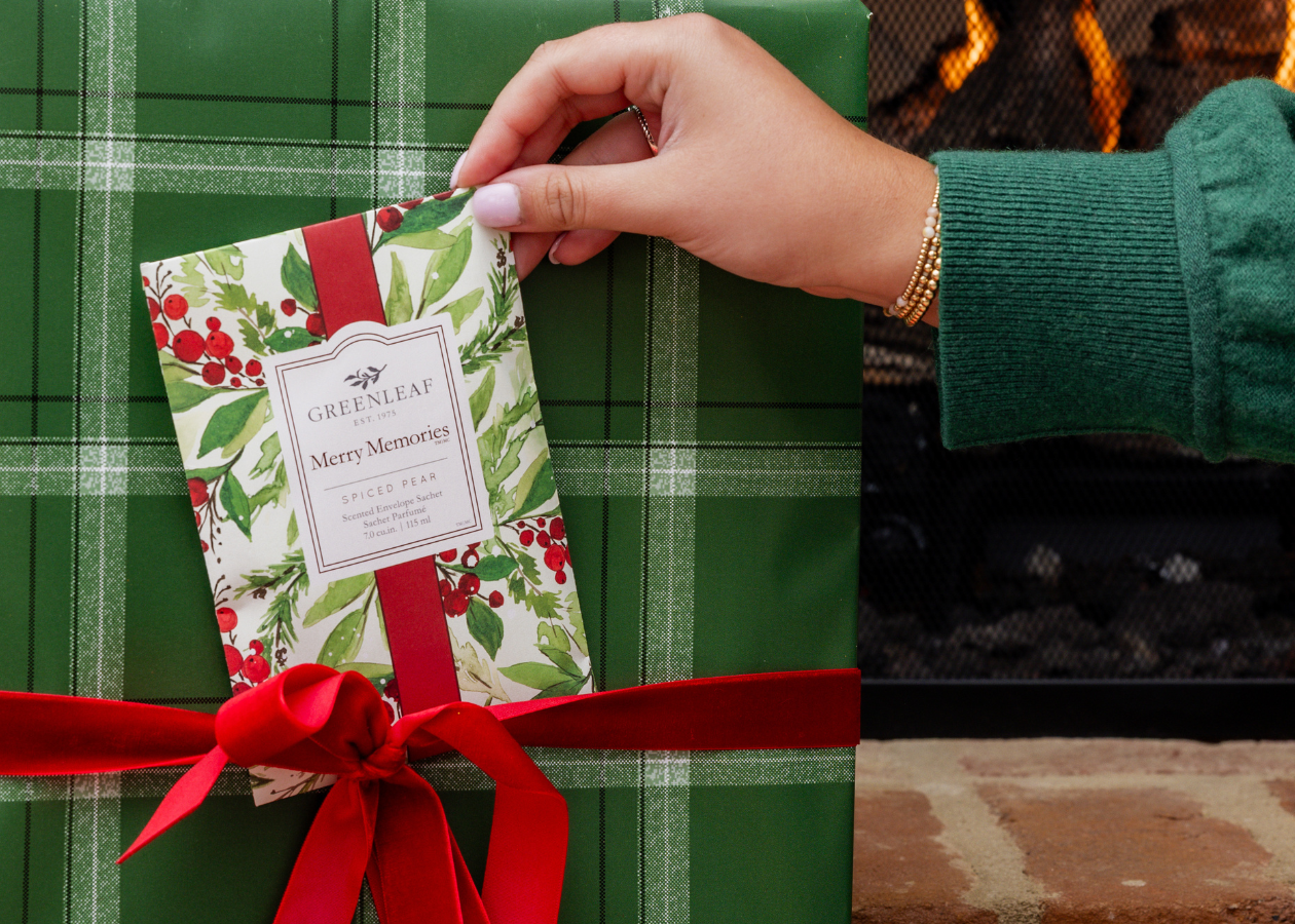 Person wrapping a gift with a green plaid paper and red ribbon, with a fireplace and Christmas tree in the background.
