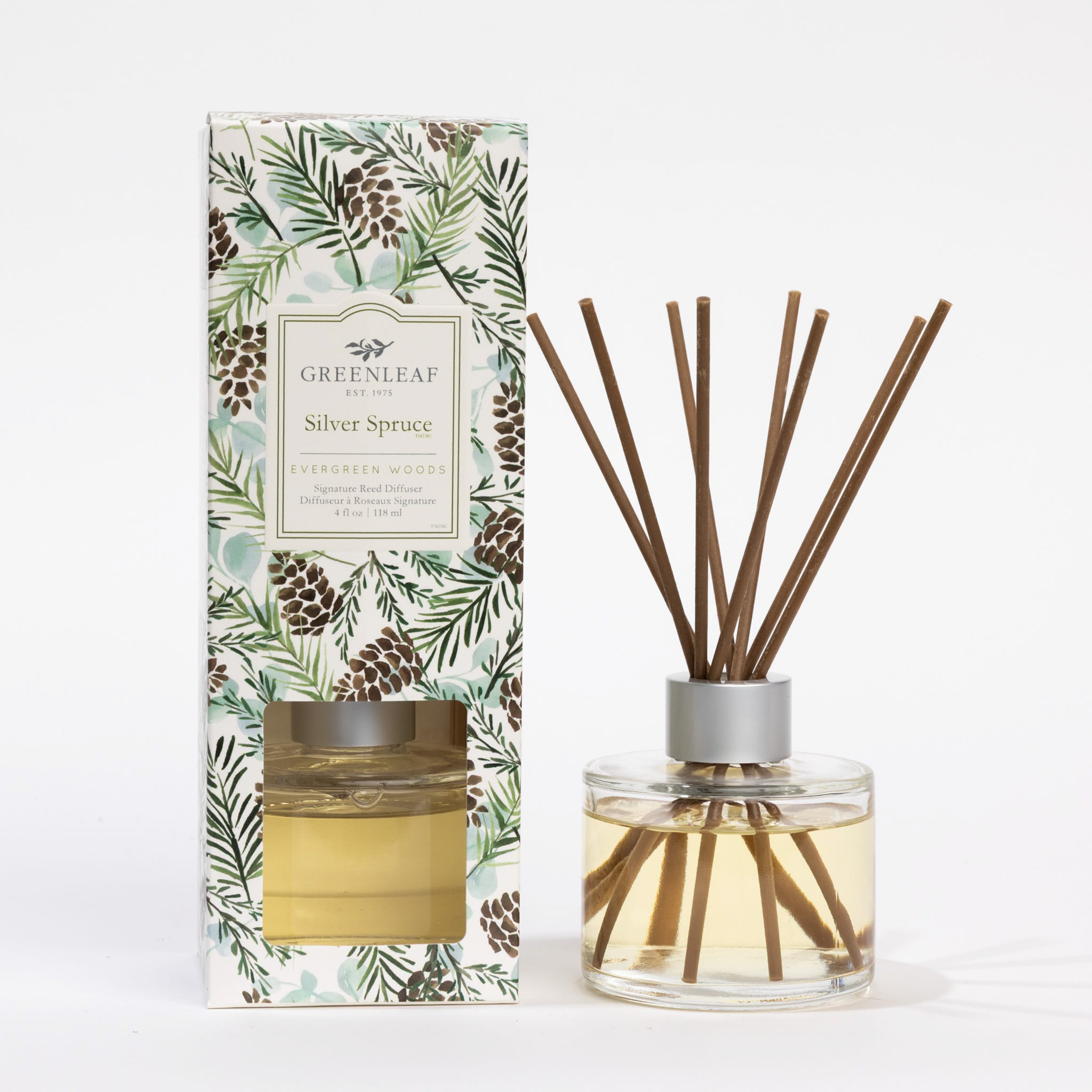 A bottle of Greenleaf Gifts Silver Spruce Reed Diffuser with brown fiber reeds stands beside its decorative box with pinecones and green foliage. The label reads Evergreen Woods, reflecting the Silver Spruce scent inside.