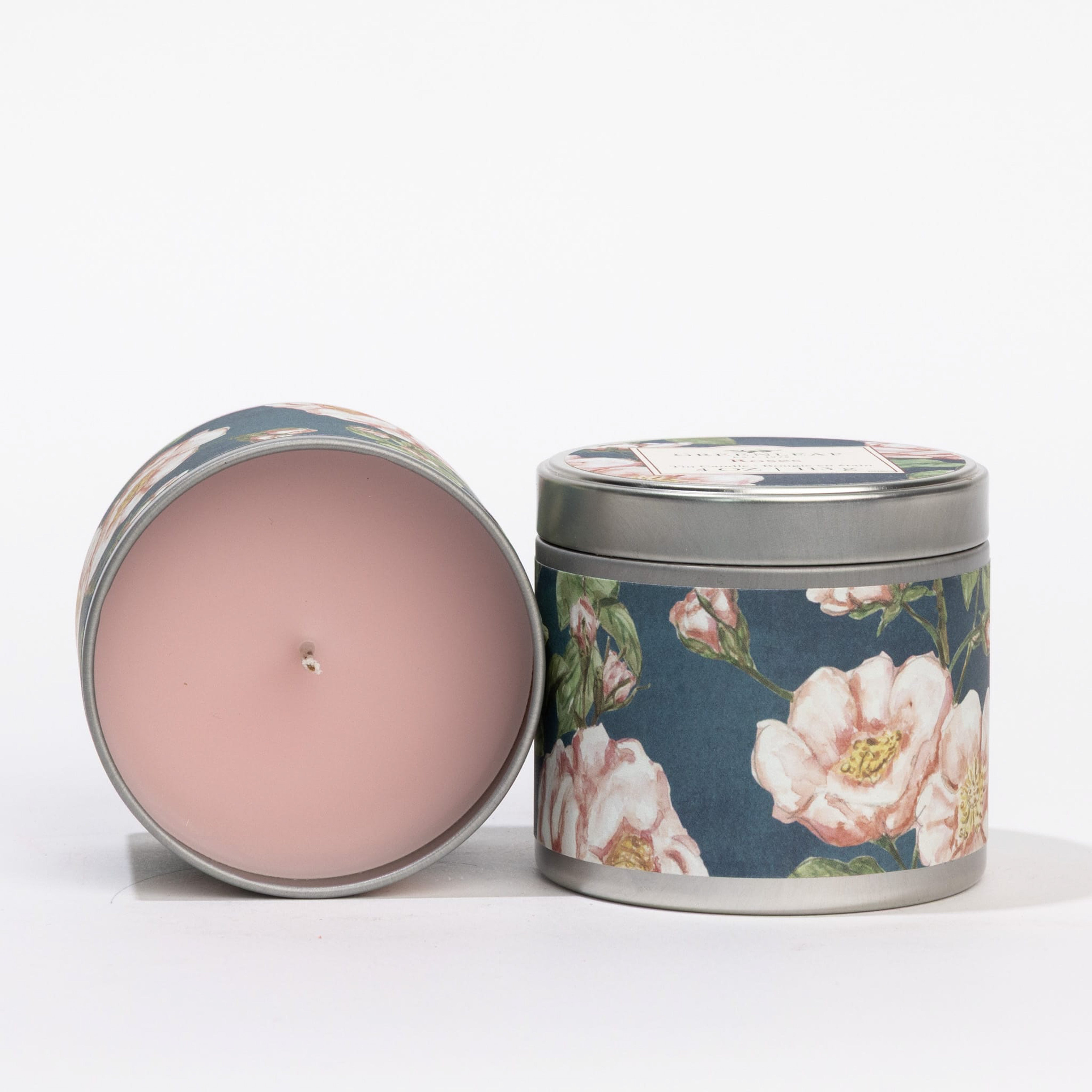 The Roses Tin Candle by Greenleaf Gifts features two decorative tins with pink wax and a rose scent. One stands upright in a floral dark blue tin; the other lies on its side revealing the pink wax and wick, both displayed on a white surface.