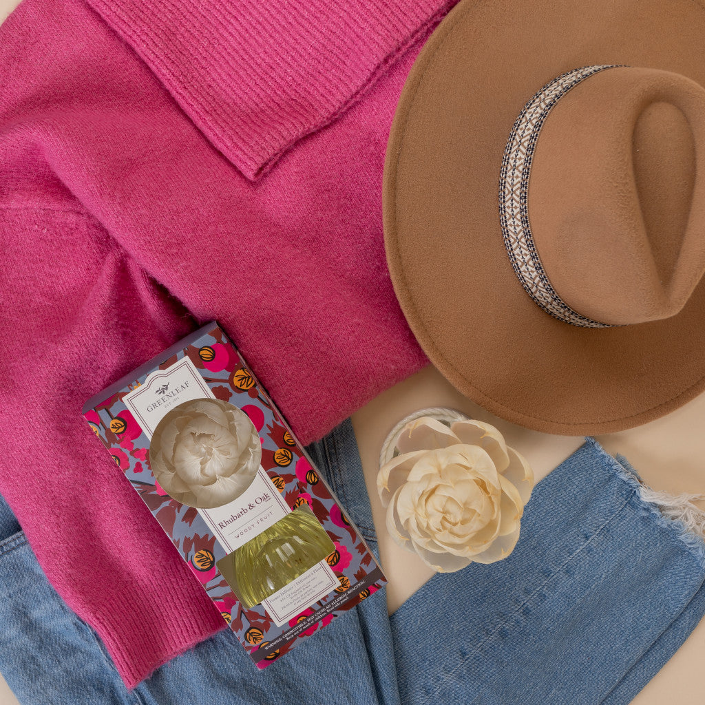 A flat lay features a pink sweater, brown wide-brim hat with decorative band, blue jeans, Greenleaf Gifts’ Rhubarb & Oak Flower Diffuser (boxed), and a cream artificial flower on a beige background.