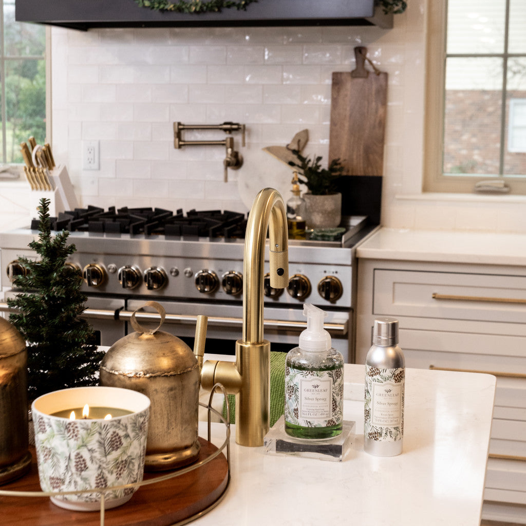A modern kitchen features a gold faucet and the Silver Spruce Patterned 3 Wick Candle by Greenleaf Gifts on a white island. A small Christmas tree and festive decor enhance the bright space, with a stainless steel stove in the background.