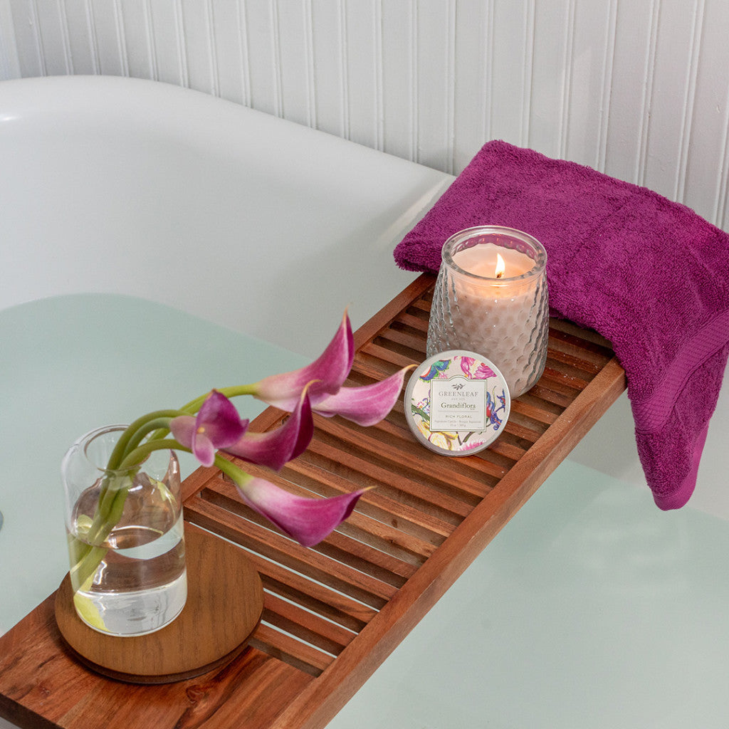 A Grandiflora scented candle on a tray across a bath rub with a flower and hand towel