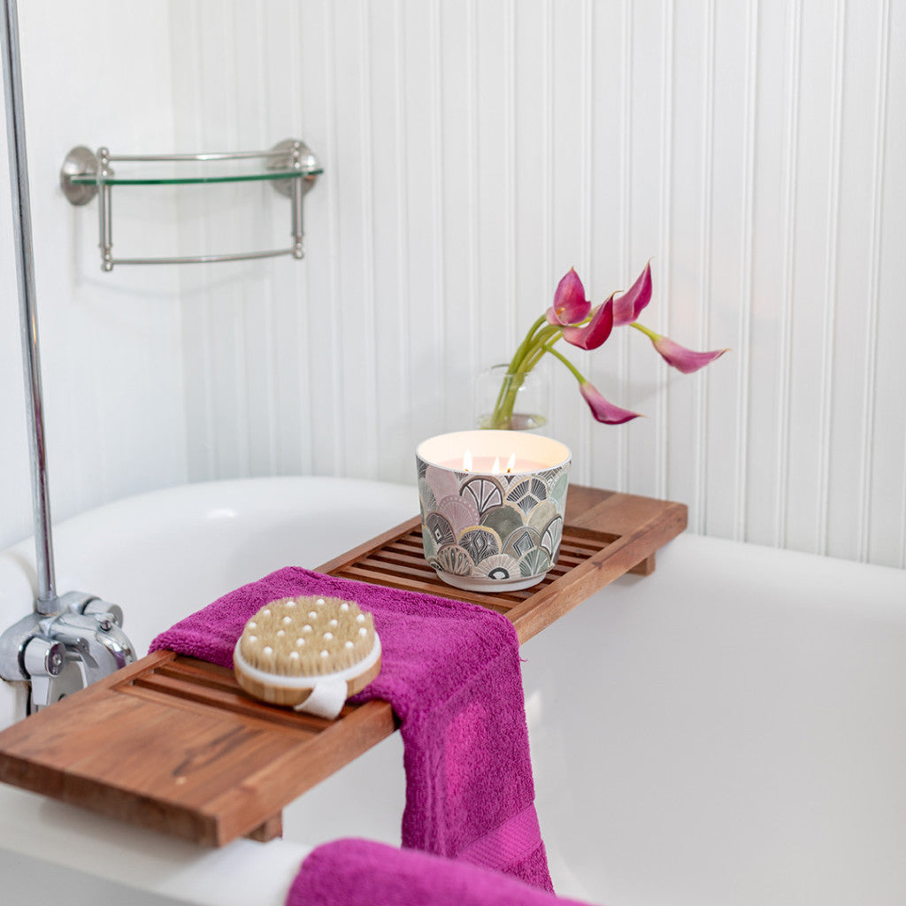 A white bathtub with a wooden tray holding Greenleaf Gifts Patterned 3 Wick Candle - Haven, a purple towel, and a bath brush. In the background, three pink calla lilies sit in a glass vase against a white paneled wall.
