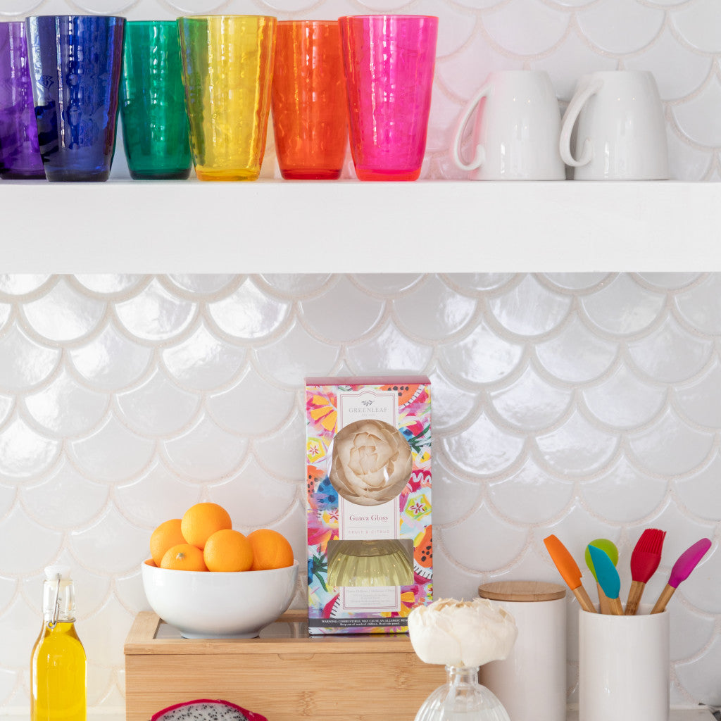 A bright kitchen shelf displays colorful glasses, white mugs, a bowl of oranges, a floral-patterned box, utensils with vibrant handles in a white jar, and the Guava Gloss Flower Diffuser by Greenleaf Gifts against a white scalloped tile backsplash.
