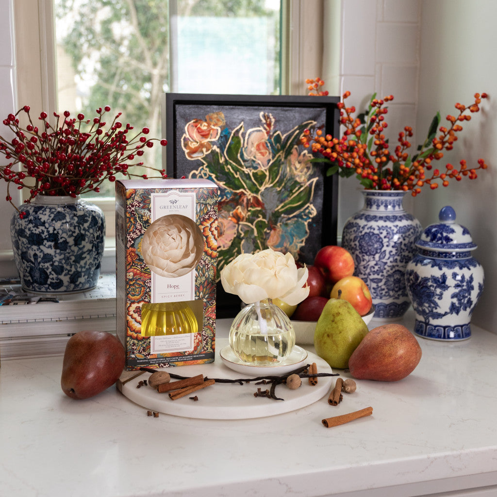 A kitchen counter features pears, apples, cinnamon sticks, and the Hope Flower Diffuser by Greenleaf Gifts. Blue and white vases with red berries, a floral painting, and a window appear in the background.
