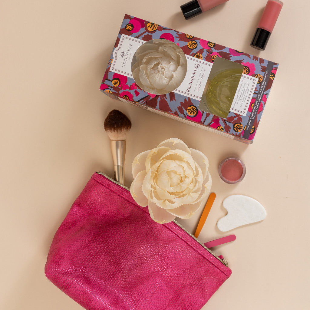 A pink makeup bag with brushes, a gua sha stone, lip balm, and a white artificial flower hint at the Rhubarb & Oak Flower Diffuser by Greenleaf Gifts in a colorful box. Two lip products rest nearby on the beige surface.