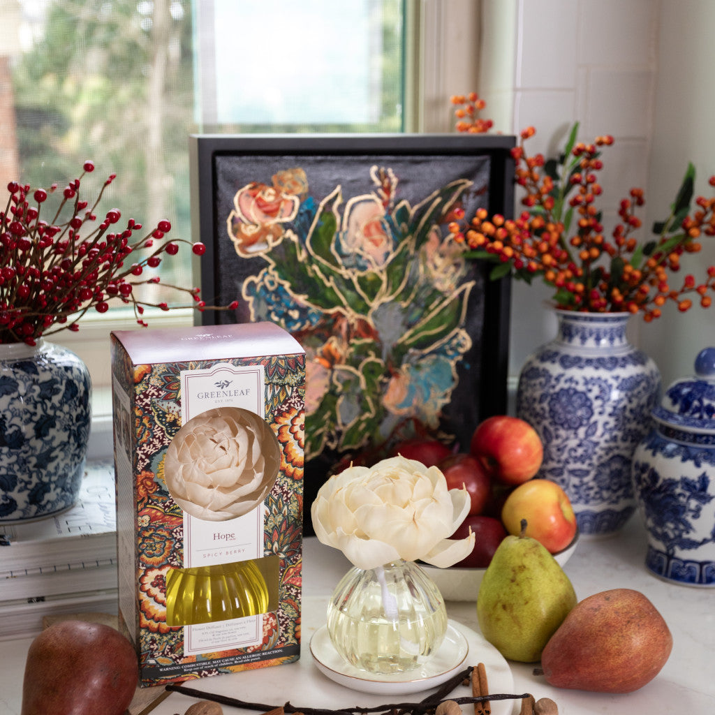 The Hope Flower Diffuser by Greenleaf Gifts, with its elegant white bloom and ornate box, sits on a table amid pears, apples, vases with red berries, and a floral painting—ready to infuse your space with your favorite scent.