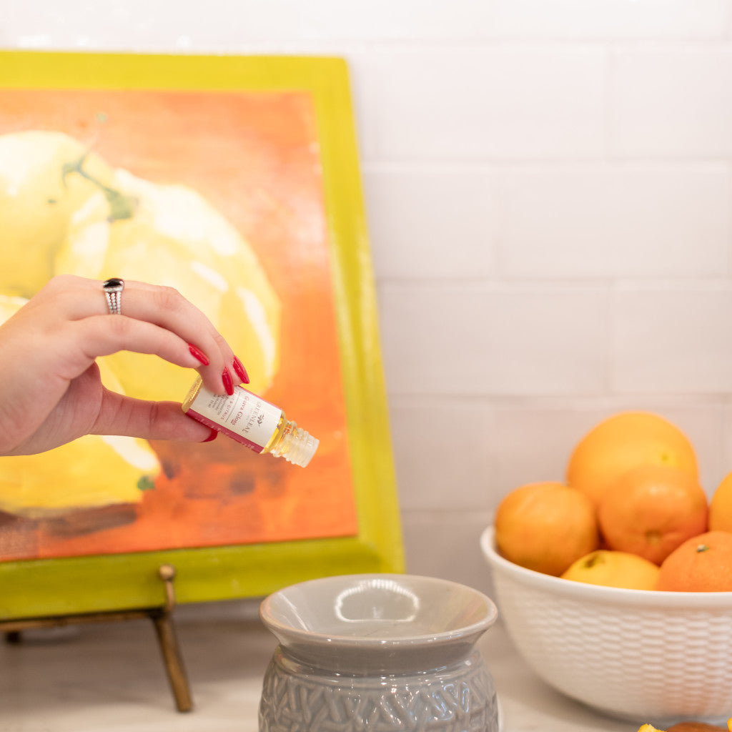 A hand with red nail polish pours Greenleaf Gifts Guava Gloss Home Fragrance Oil into a gray ceramic diffuser. In the background, a bowl of oranges and a painting of yellow fruit add a bright touch to the kitchen’s flameless fragrance.