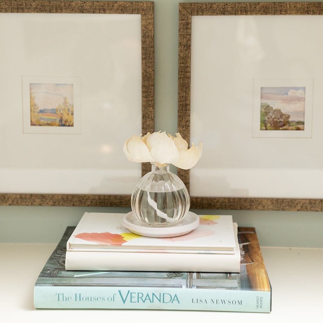 A Greenleaf Gifts Flower Diffuser-Magnolia (GLG966515S), featuring white blossoms in an 8 oz glass vase, sits atop a floral-covered book and The Houses of Veranda by Lisa Newsom, in front of two framed landscape prints.