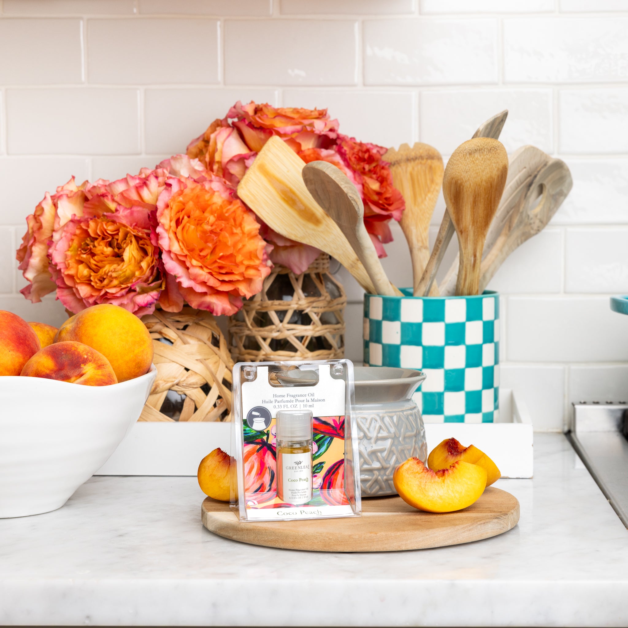 A small bottle of Greenleaf Gifts' Coco Peach Home Fragrance Oil sits on a wooden tray with peach slices. Behind it are fresh peaches in a white bowl, floral decor, and utensils in a checkered holder on the kitchen counter.