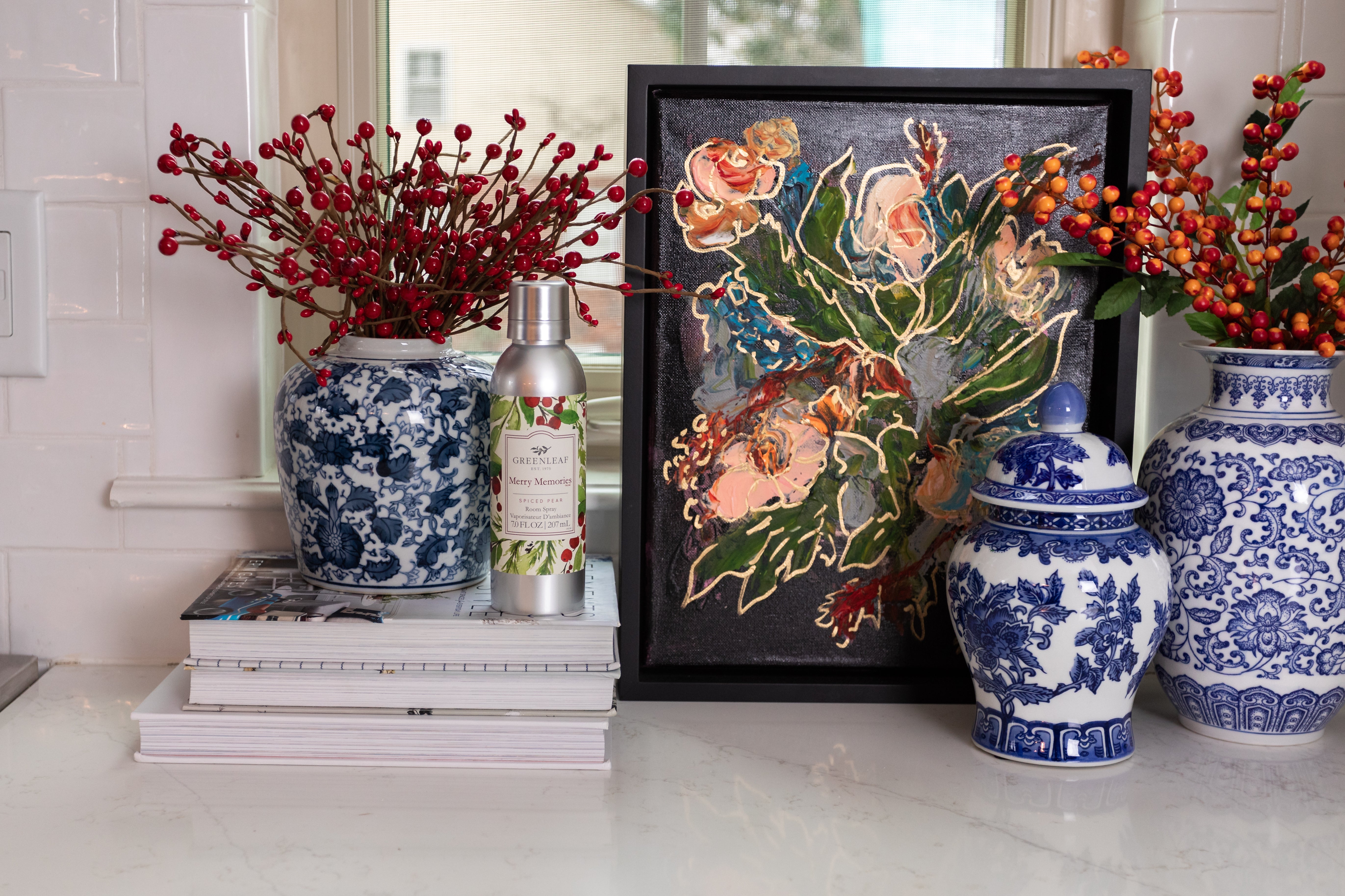A countertop with blue and white vases, red and orange berry branches, stacked books, framed floral art, and a Greenleaf Gifts Merry Memories Room Spray sits before a white tiled backsplash and a window.