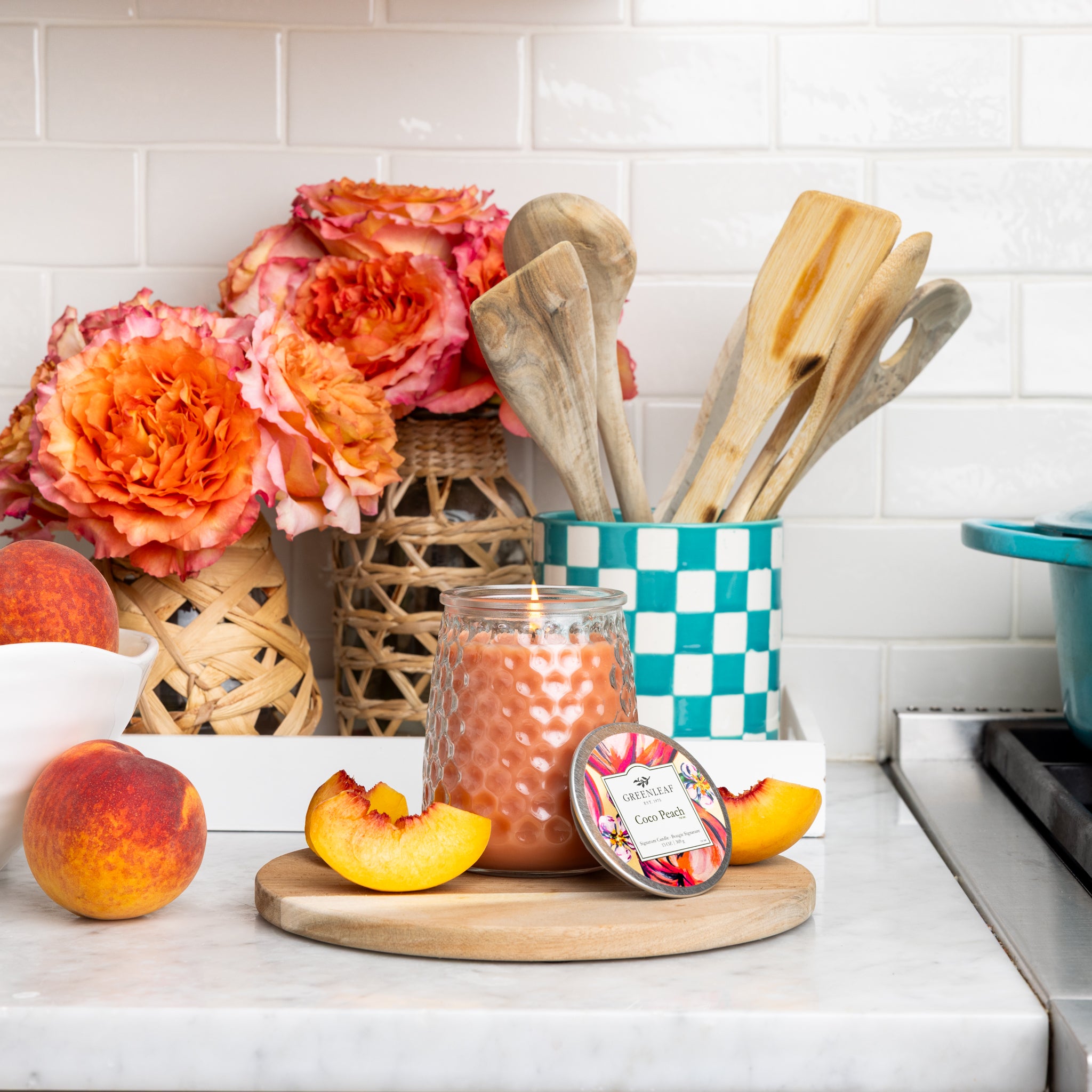 A Greenleaf Gifts Coco Peach Signature Candle with a metal lid rests on a wooden board beside fresh peaches and peach slices, accented by orange-pink roses in a woven vase and kitchen utensils in a blue checkered holder. Enjoy Greenleaf fragrances.