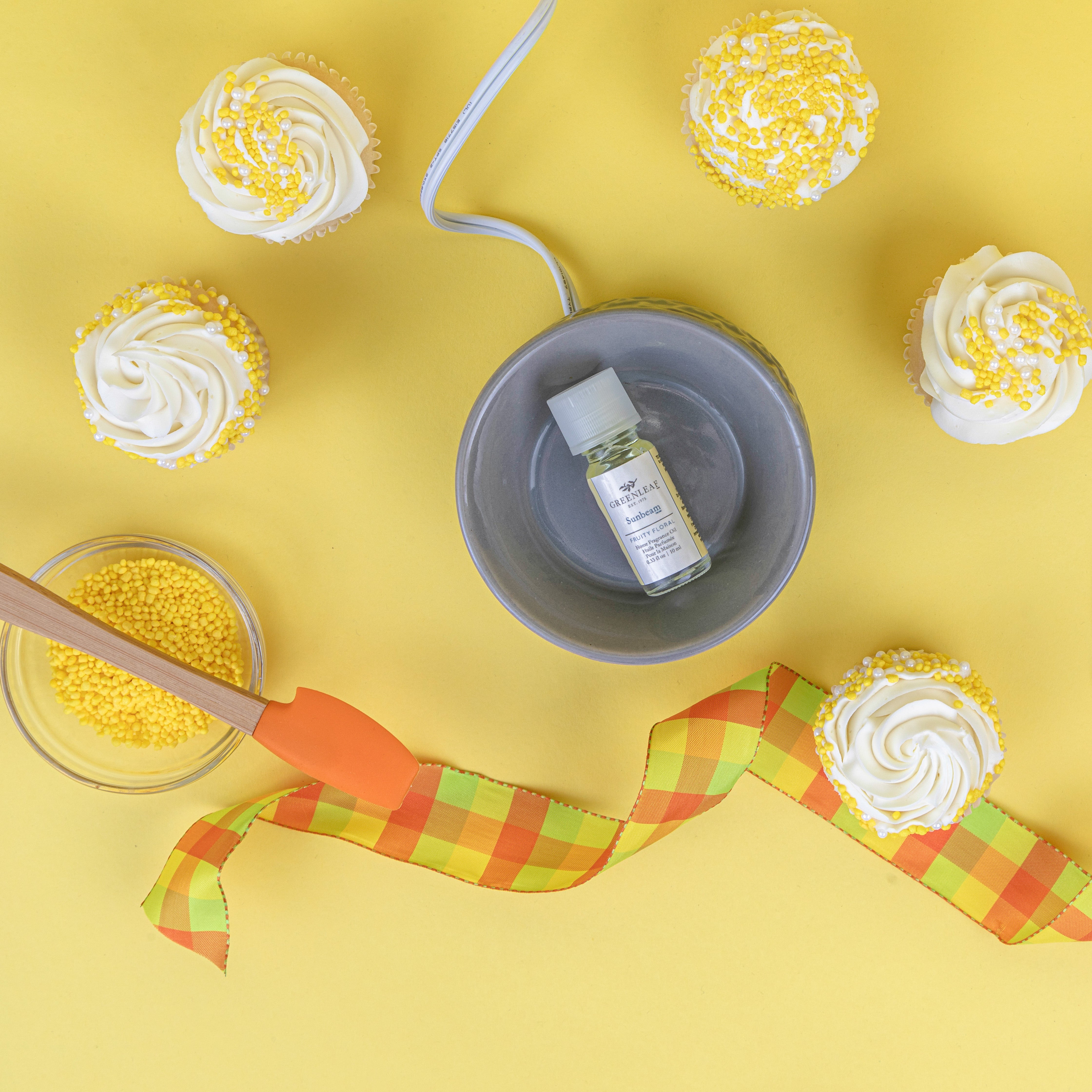 A flat lay featuring cupcakes with white frosting and yellow sprinkles, a dish of yellow sprinkles with an orange spatula, a Greenleaf Gifts Sunbeam Home Fragrance Oil bottle in a bowl, and a plaid ribbon on a vibrant yellow background.