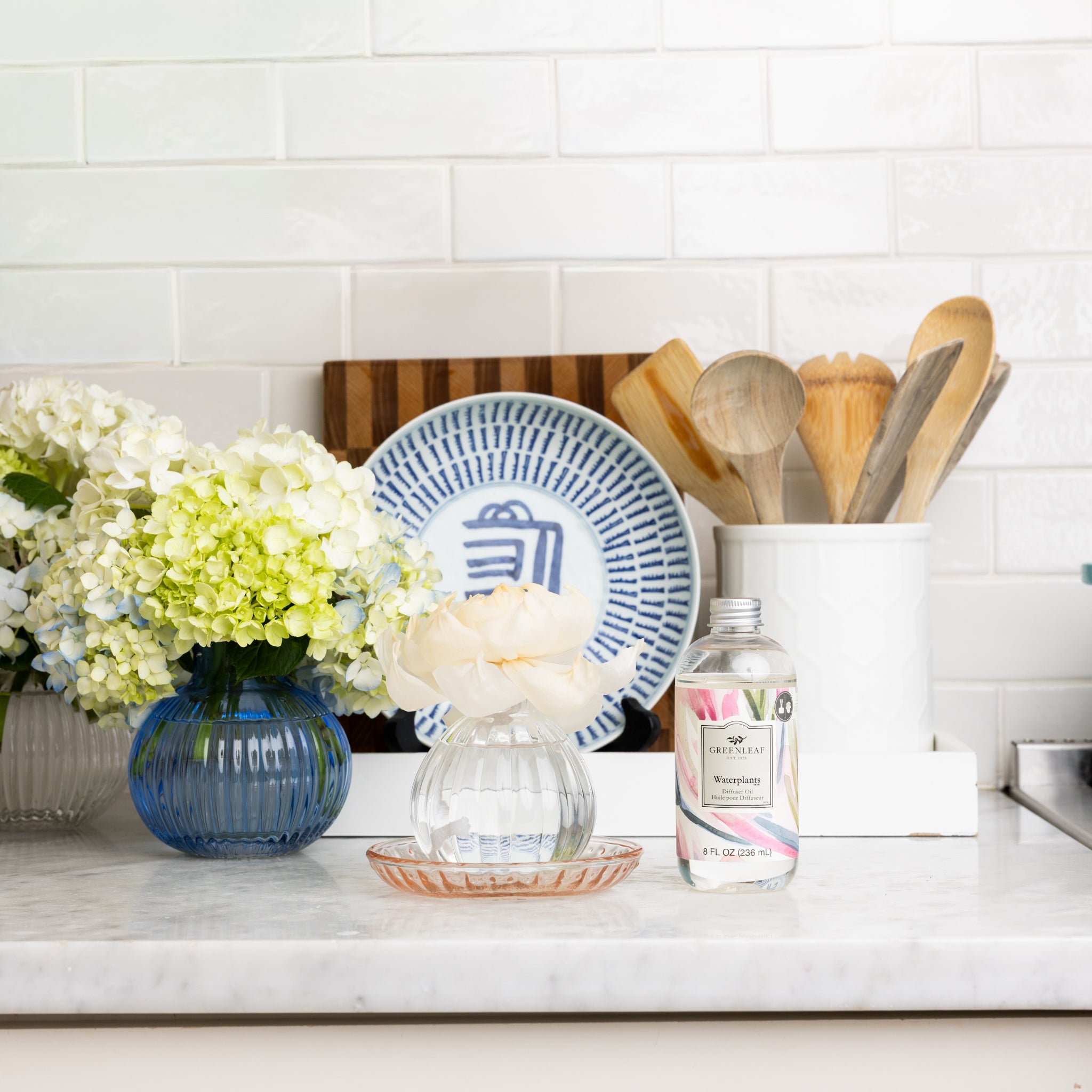 A kitchen counter with blue and white flowers in vases, a bottle of rose water, Greenleaf Gifts Waterplants Diffuser Oil, a glass dish, wooden utensils in a white holder, and blue and white plates against a white subway tile backsplash.