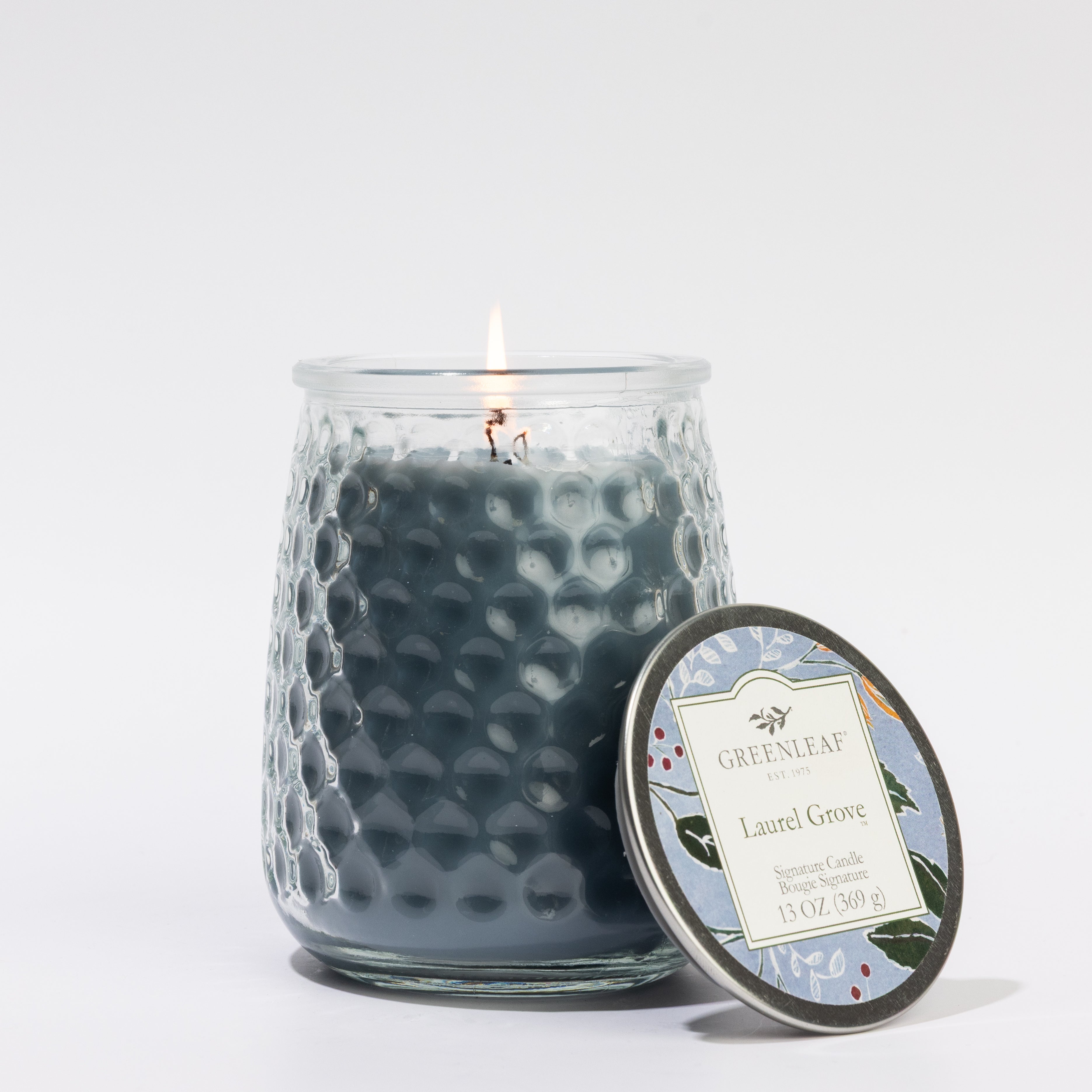 A lit dark blue 13oz Laurel Grove Signature Candle by Greenleaf Gifts sits in a textured glass jar with its metal lid beside it and label visible; lasts 90–100 hours.