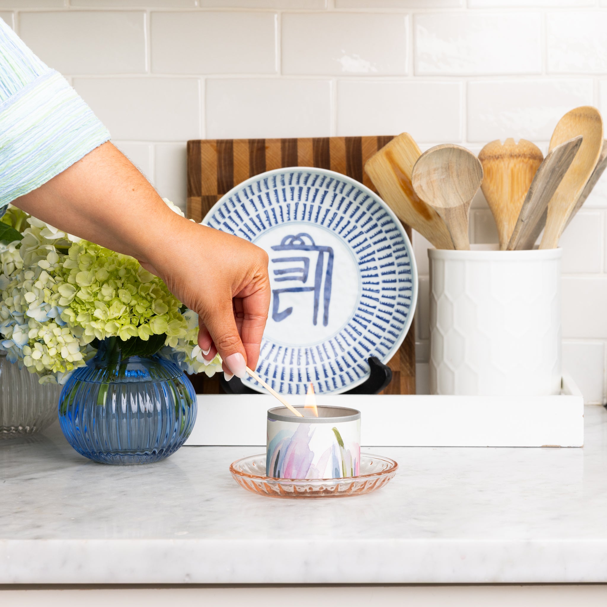 A hand lights the Greenleaf Gifts Waterplants Tin Candle in a decorative holder on a marble kitchen counter, beside a blue vase of flowers, a patterned plate, and a white holder with wooden spoons.