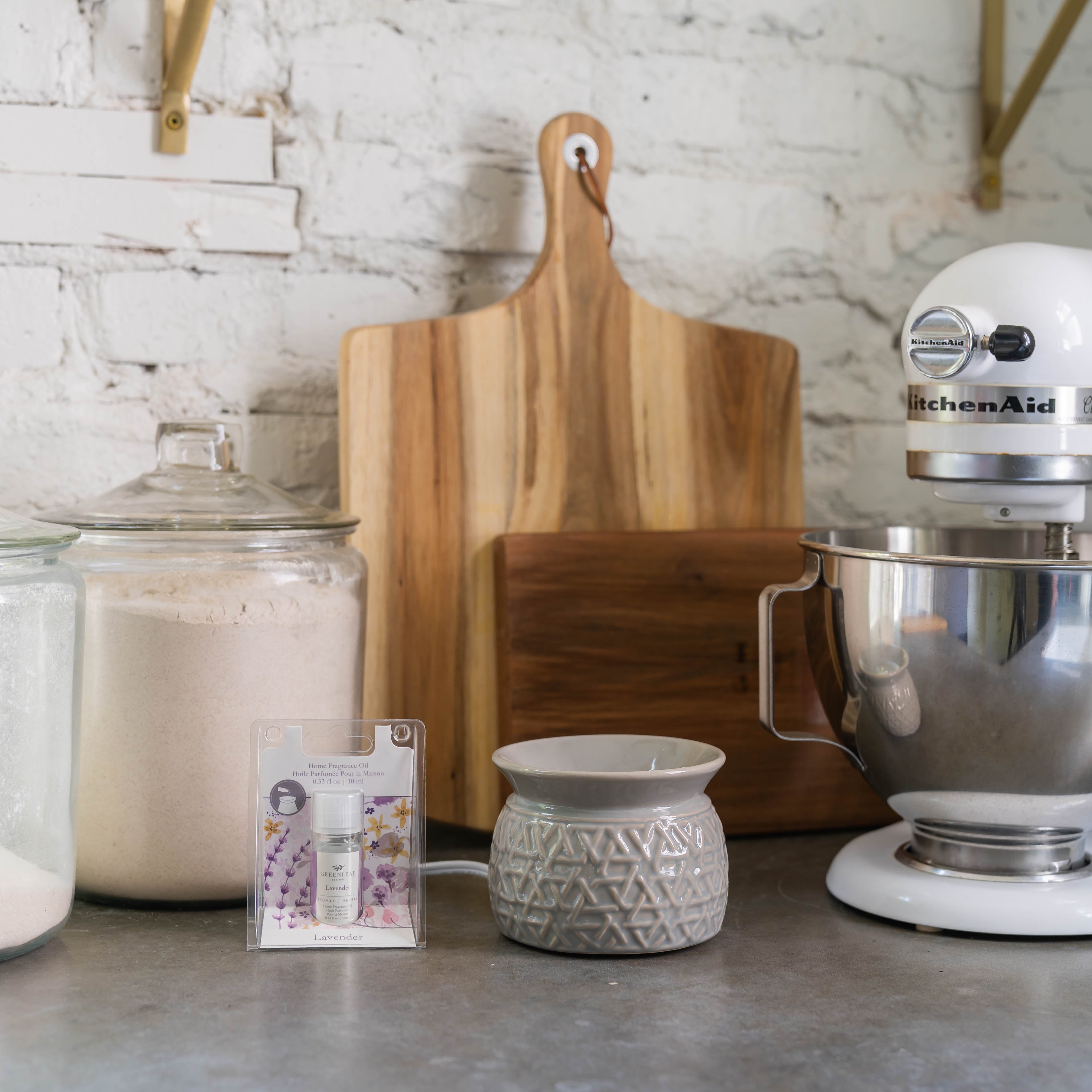 On the kitchen counter are glass jars of flour and sugar, a wooden cutting board, a gray ceramic fragrance warmer, a white KitchenAid stand mixer, and Greenleaf Gifts Lavender Home Fragrance Oil.
