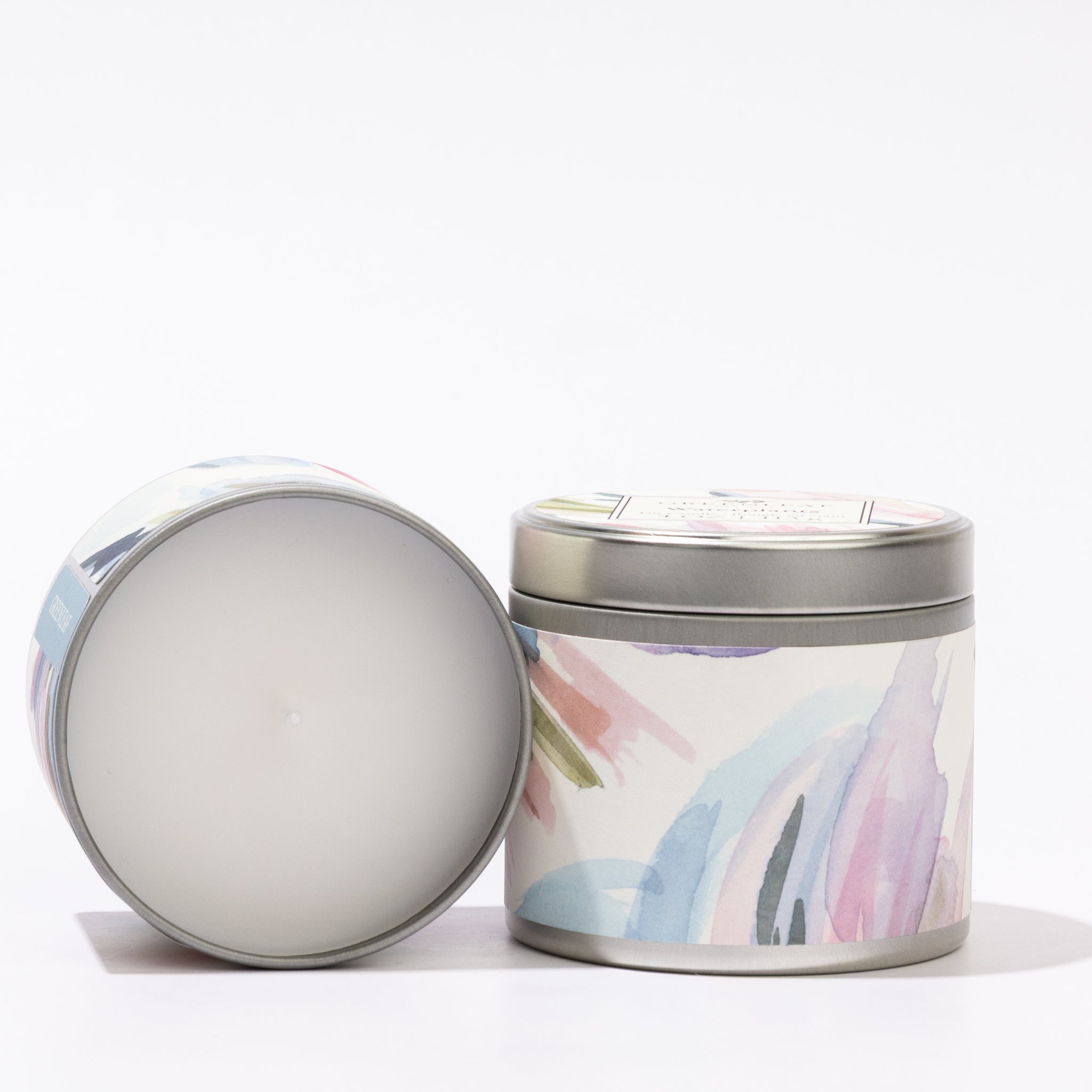 The Greenleaf Gifts Waterplants Tin Candle features abstract pastel designs on silver tins; one stands upright with a lid, the other displays white wax—perfect as a portable candle with a calming waterplants scent.