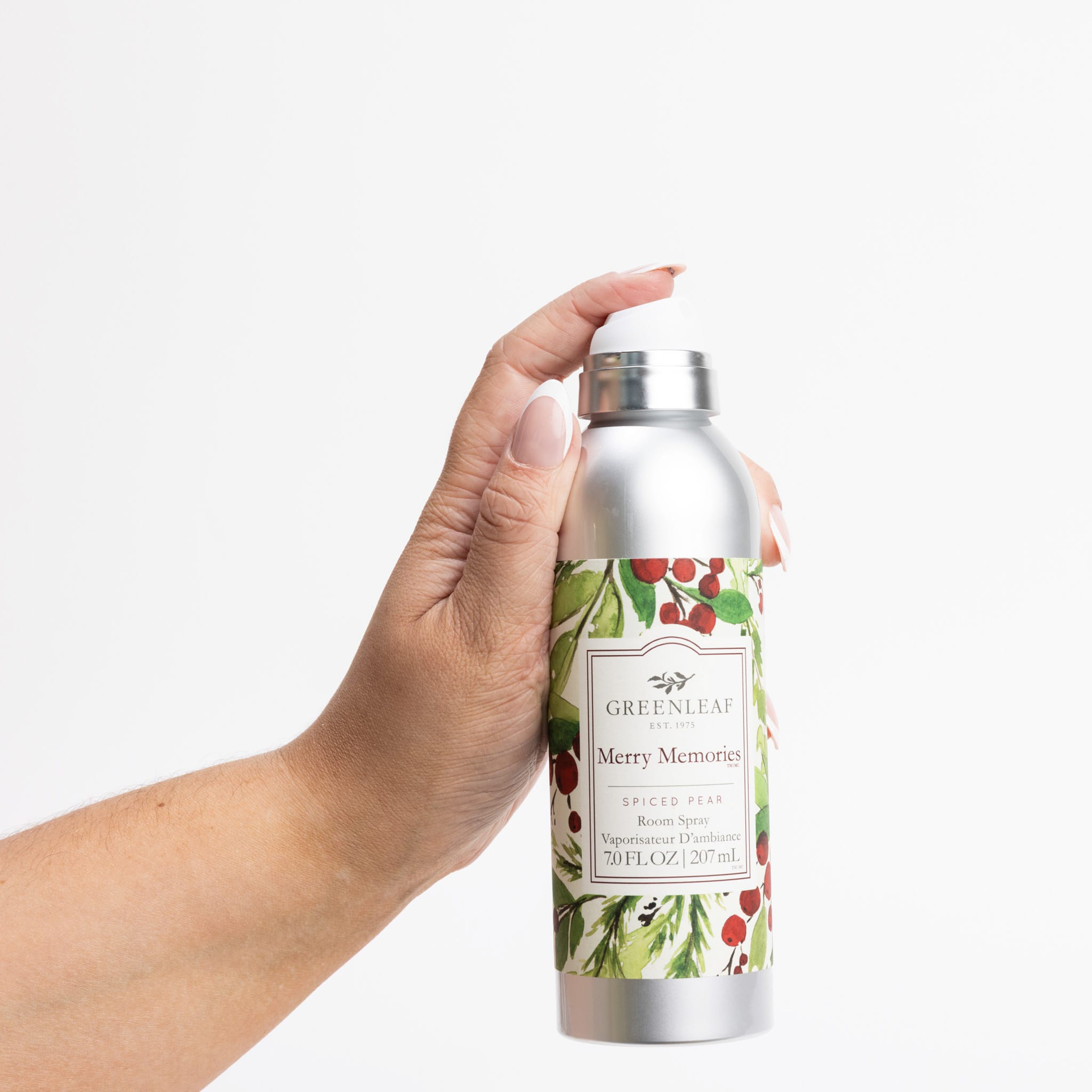 A manicured hand holds a silver bottle labeled Greenleaf Gifts Merry Memories Room Spray, adorned with festive red berries and green leaves, set against a white background.