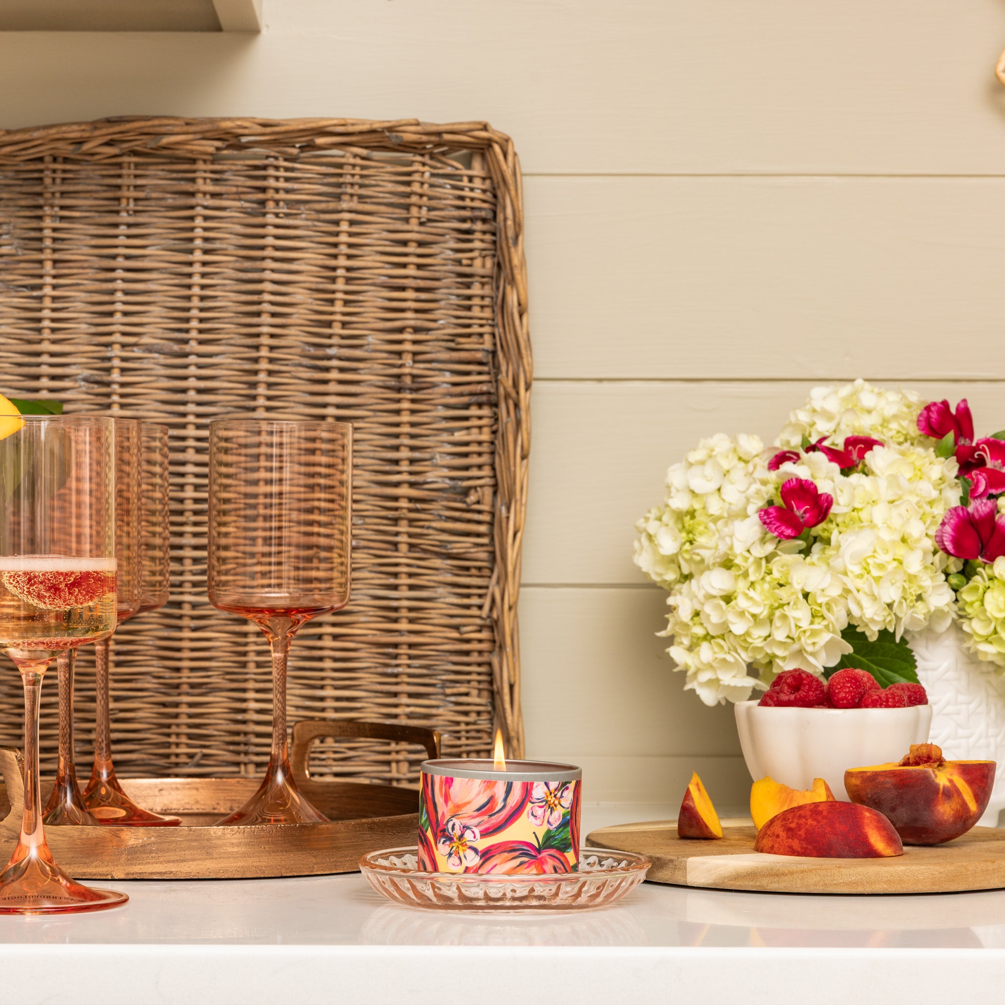 On a kitchen counter against a light wall sit pink wine glasses, a Greenleaf Gifts Coco Peach Tin Candle, a glass plate, white hydrangeas with red blooms in a vase, strawberries, sliced peaches in a bowl, and a wicker tray.