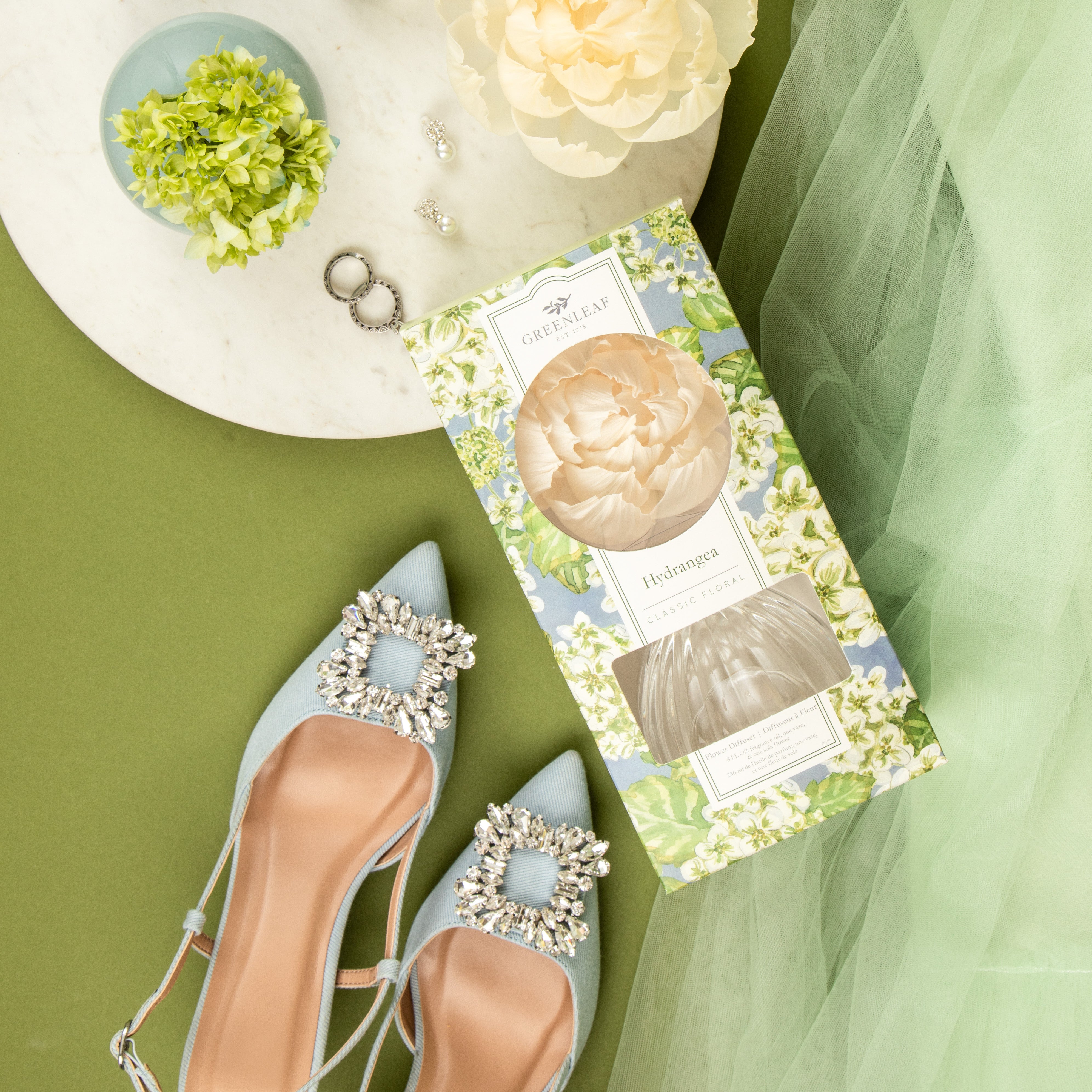 Flat lay of elegant blue heels with rhinestone buckles, a Greenleaf Gifts Hydrangea Flower Diffuser, a sola paper flower, green hydrangea, green tulle fabric, earrings, and rings arranged on a round white tray.