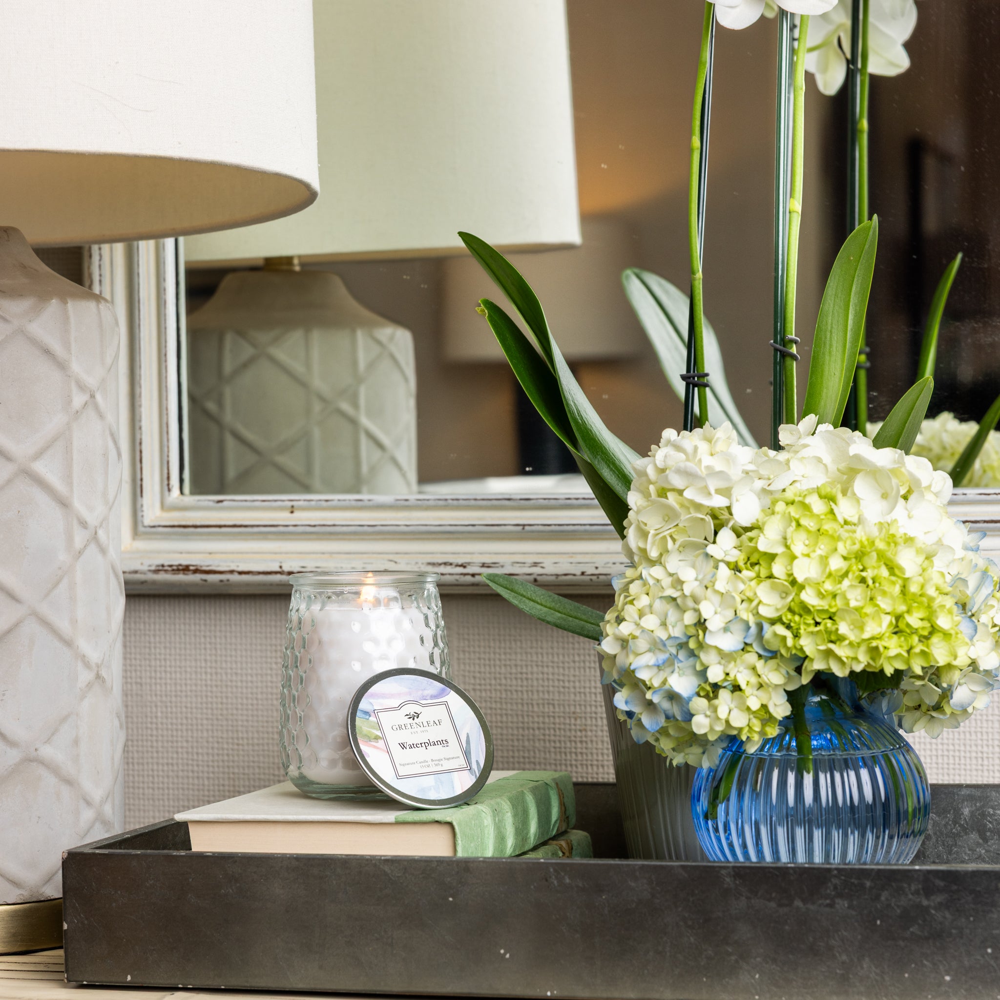 A tray holds a vase with white and green hydrangeas, a green book, a white book, and a lit Greenleaf Gifts Waterplants Signature Candle in a glass jar. In the background, a lamp and mirror reflect part of the peaceful setting.